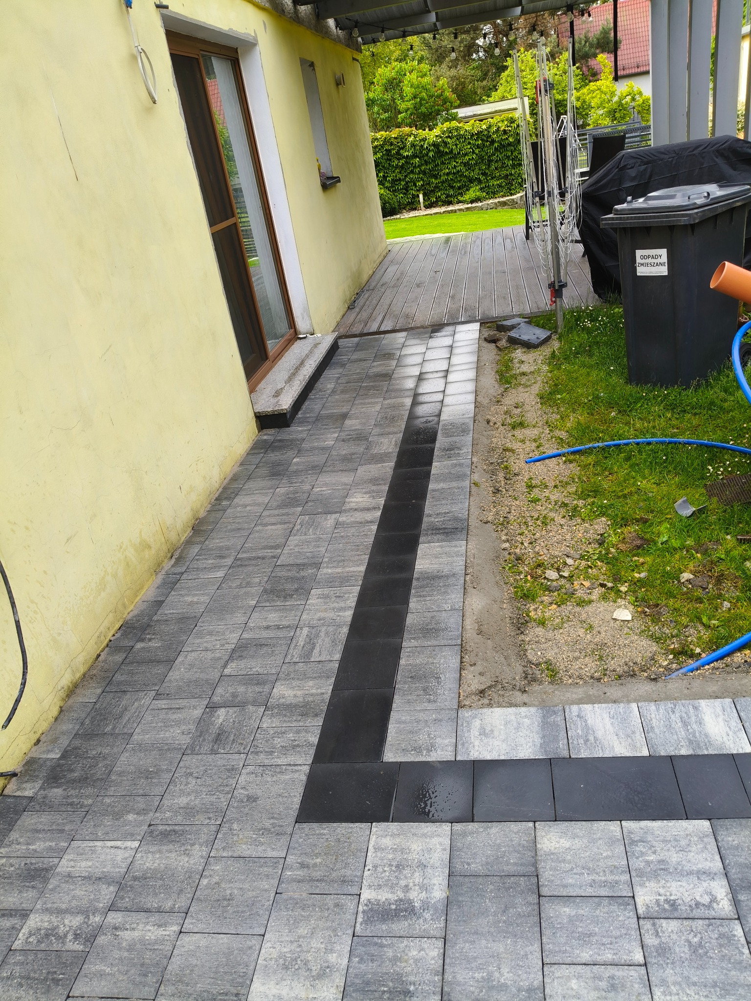 Ułożona kostka brukowa w odcieniach szarości z czarnym pasem, prowadząca od drzwi domu do trawnika. Widoczny fragment elewacji budynku i elementy otoczenia.