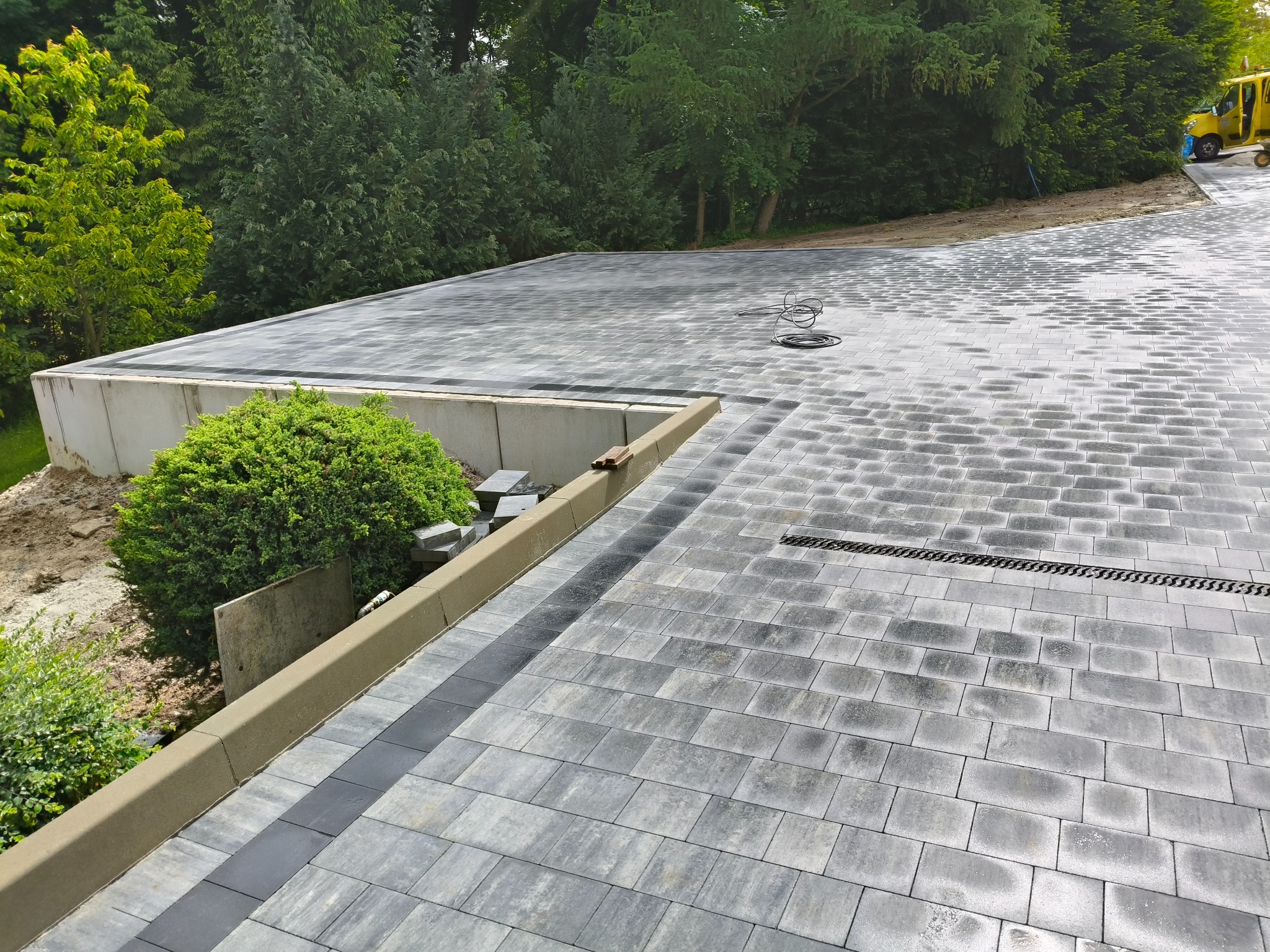 Świeżo ułożona, szara kostka brukowa z odpływem liniowym i obrzeżami, otoczona zielenią. Widoczny żółty pojazd. Układanie kostki z dbałością o detale.