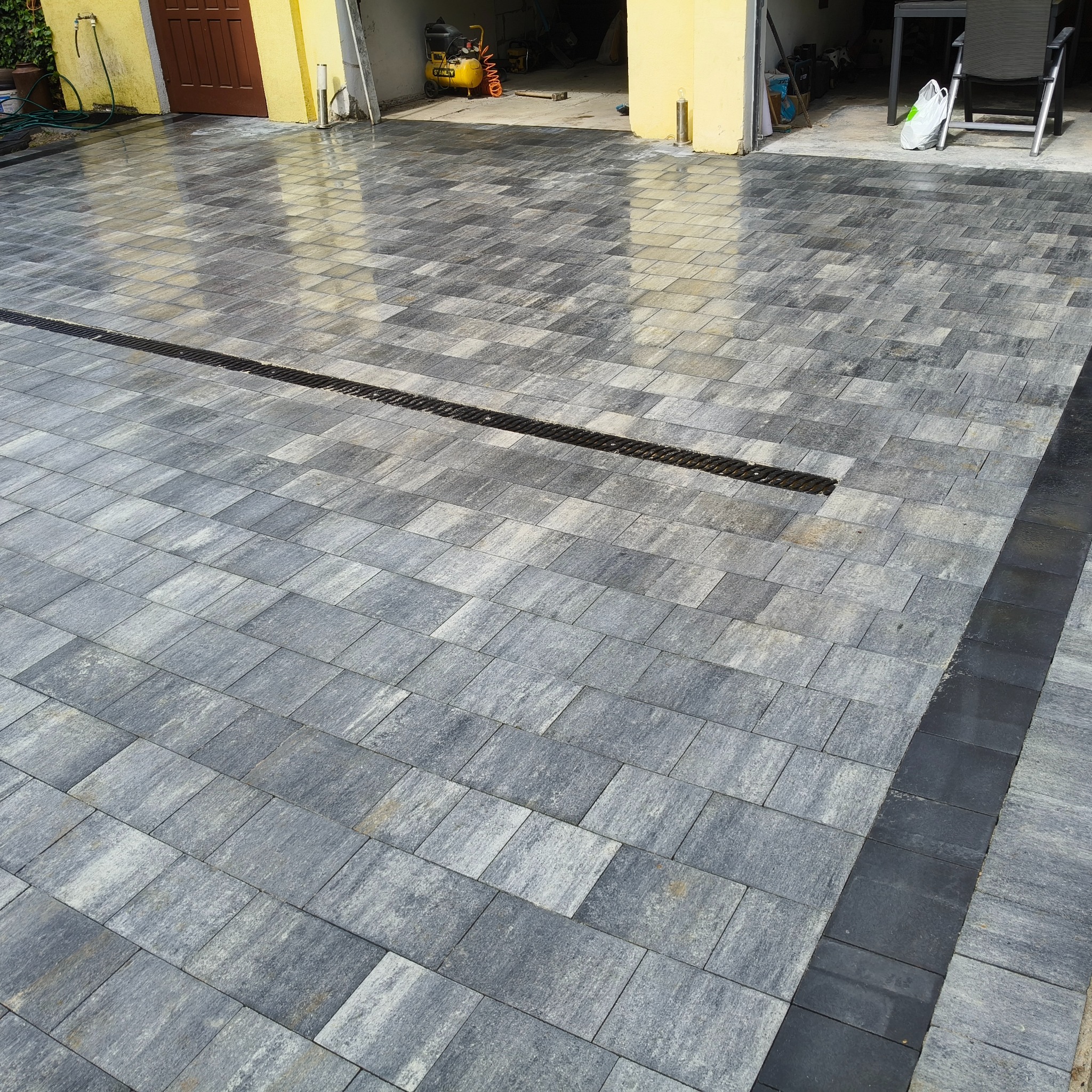 Ułożona szara kostka brukowa z czarnym obrzeżem i liniowym odpływem wody na podjeździe, widok z góry, w tle budynek z żółtą elewacją i otwartym garażem.