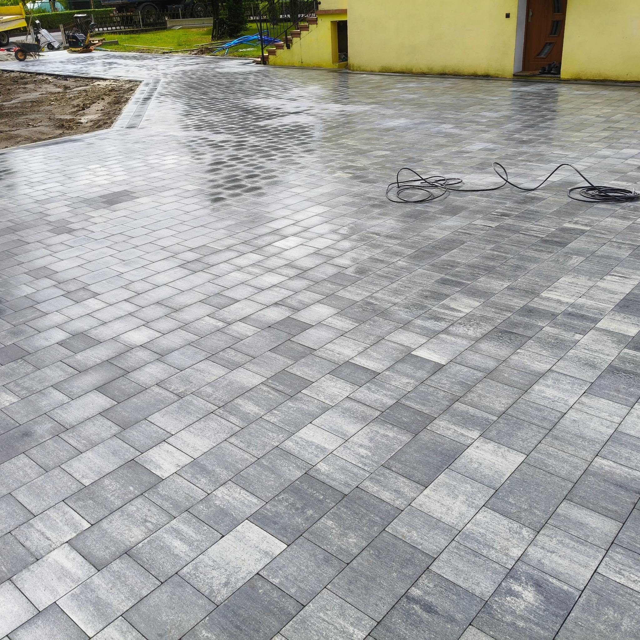 Ułożona, szara kostka brukowa tworząca dużą, lekko wilgotną powierzchnię podjazdu przed żółtym budynkiem. Widoczny kabel i fragment placu budowy.