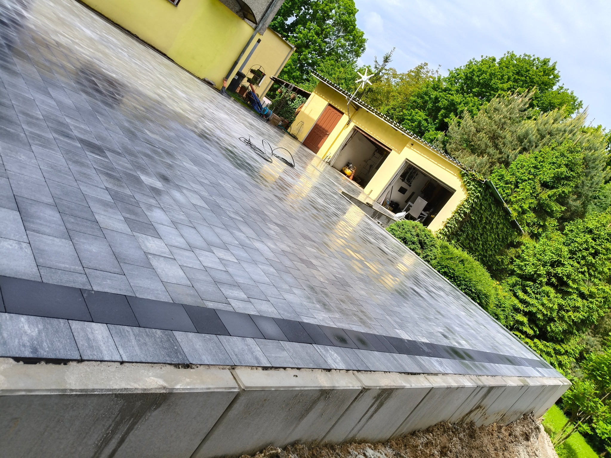 Ułożona, szara kostka brukowa z ciemniejszym pasem, obok żółty budynek z garażem i zieleń. Widok z góry, z boku, ukośnie, po deszczu.