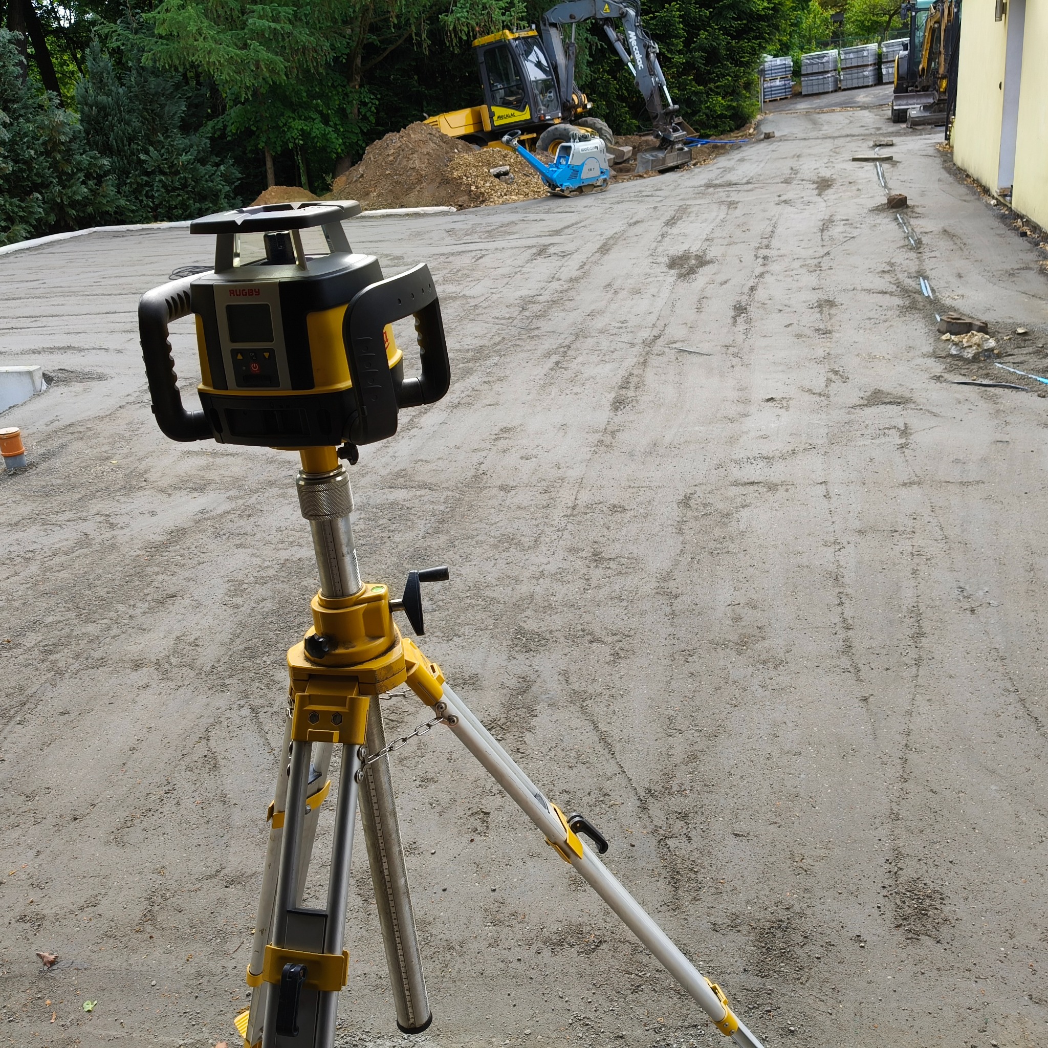 Żółty niwelator laserowy na statywie, w tle wyrównany teren pod budowę z koparką i walcem. Widoczne przygotowania pod kostkę brukową.