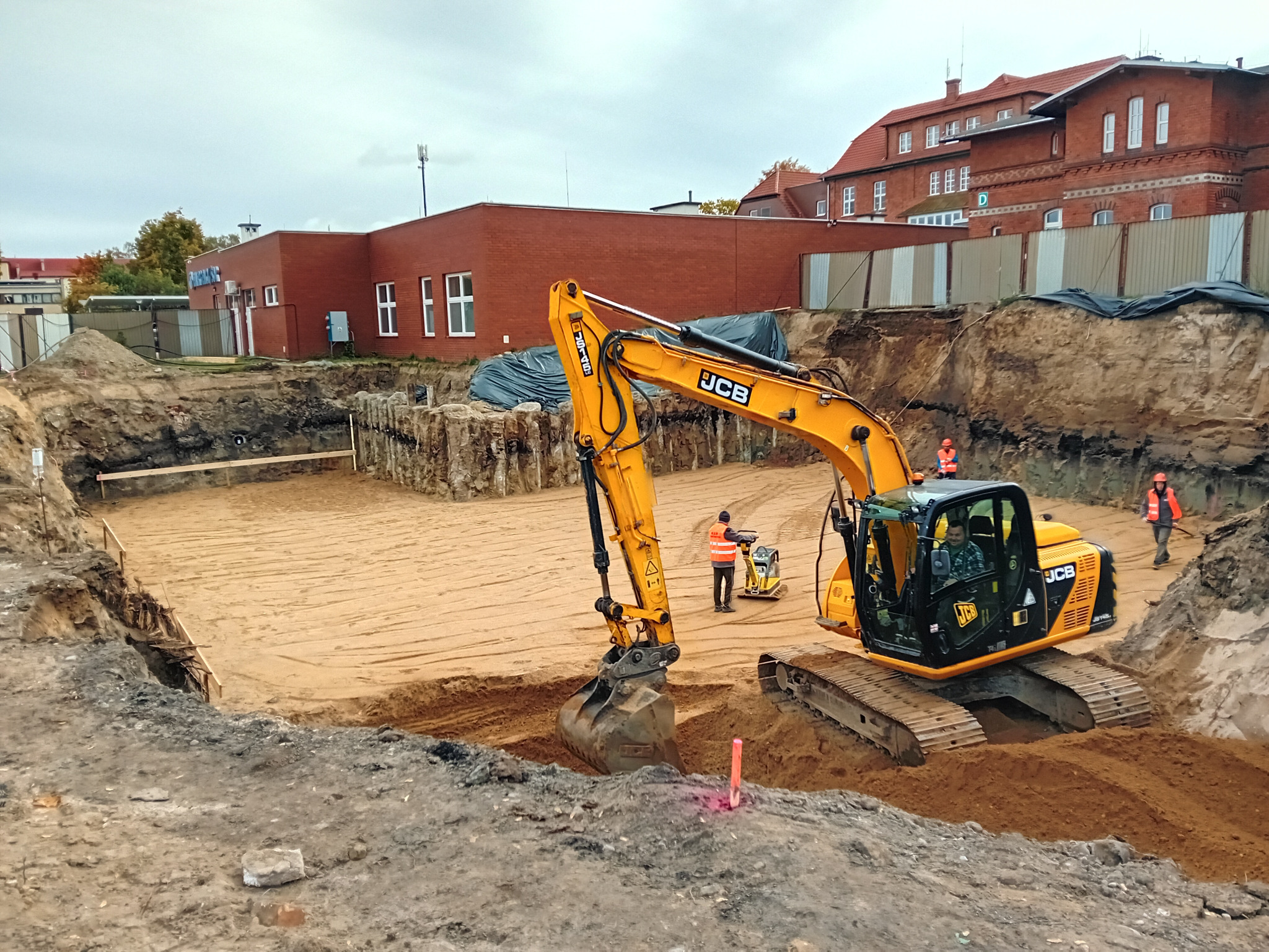 Żółta koparka JCB przy wykopie pod budowę w Giżycku. W tle budynki z czerwonej cegły i pracownicy w kamizelkach odblaskowych. Teren przygotowany pod fundamenty.