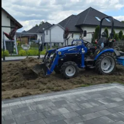 Niebieski traktor MIKANN CR-2 na działce z resztkami trawy i ziemi, przygotowujący teren pod trawnik. W tle domy i błękitne niebo z białymi chmurami.