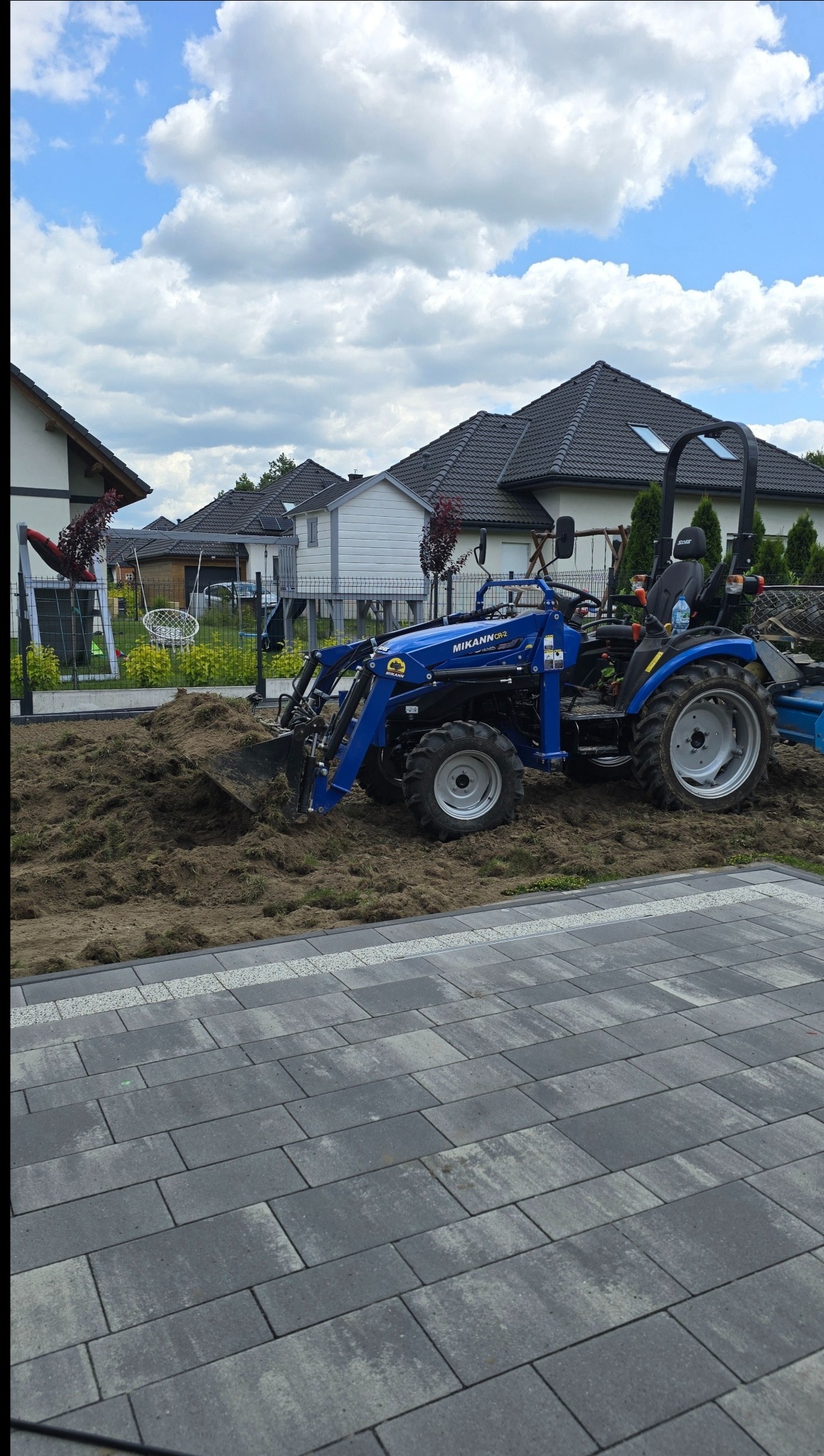 Niebieski traktor MIKANN CR-2 na działce z resztkami trawy i ziemi, przygotowujący teren pod trawnik. W tle domy i błękitne niebo z białymi chmurami.