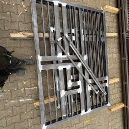 Błażej Cebula Spawart - Metalowa konstrukcja kraty, prawdopodobnie fragment bramy lub ogrodzenia, leżąca na kostce brukowej. Widoczne ślady spawania i obróbki. Elementy wsparte na drewnianych klockach.