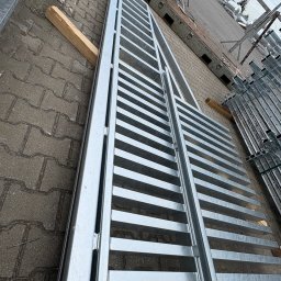 Błażej Cebula Spawart - Elementy bramy wjazdowej z metalu, przygotowane do montażu, leżą na brukowanej powierzchni. Widoczne drewniane podpórki i stosy innych elementów w tle.