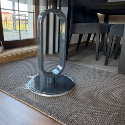 Błażej Cebula Spawart - Stół z czarnym, błyszczącym blatem i oryginalną, metalową nogą w kształcie owalu. W tle krzesła i okno. Widoczny fragment dywanu i podłogi.