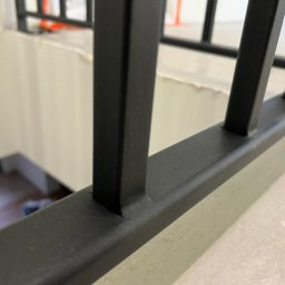 Błażej Cebula Spawart - Czarne, metalowe balustrady schodowe z bliska, widoczne pionowe i poziome elementy konstrukcji. W tle jasne ściany i schody w dół. Minimalistyczny design.