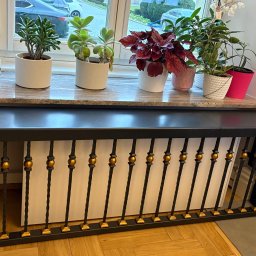 Błażej Cebula Spawart - Dekoracyjny grzejnik z metalową balustradą w kolorze czarnym i złotym, na parapecie okna ustawione doniczki z roślinami. Nietypowe rozwiązanie aranżacyjne.