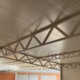 Błażej Cebula Spawart - Metalowa konstrukcja zadaszenia garażu z blachy trapezowej, widok od wewnątrz, z fragmentem ceglanego budynku w tle. Jasne oświetlenie.
