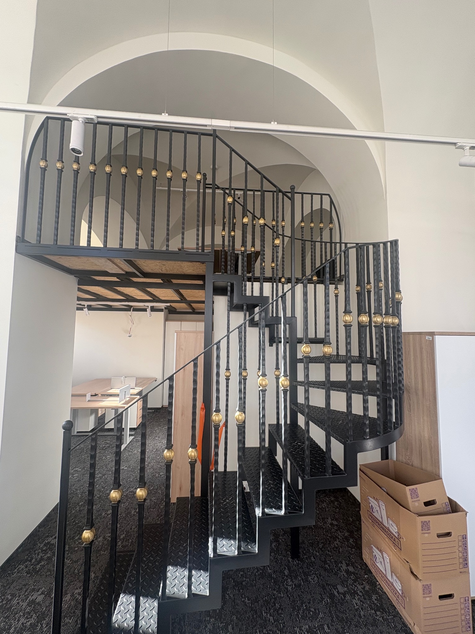 Spiralne schody metalowe z czarną konstrukcją i złotymi zdobieniami w Popowicach. Widok z dołu, ukazujący detale balustrady i ażurową konstrukcję schodów.