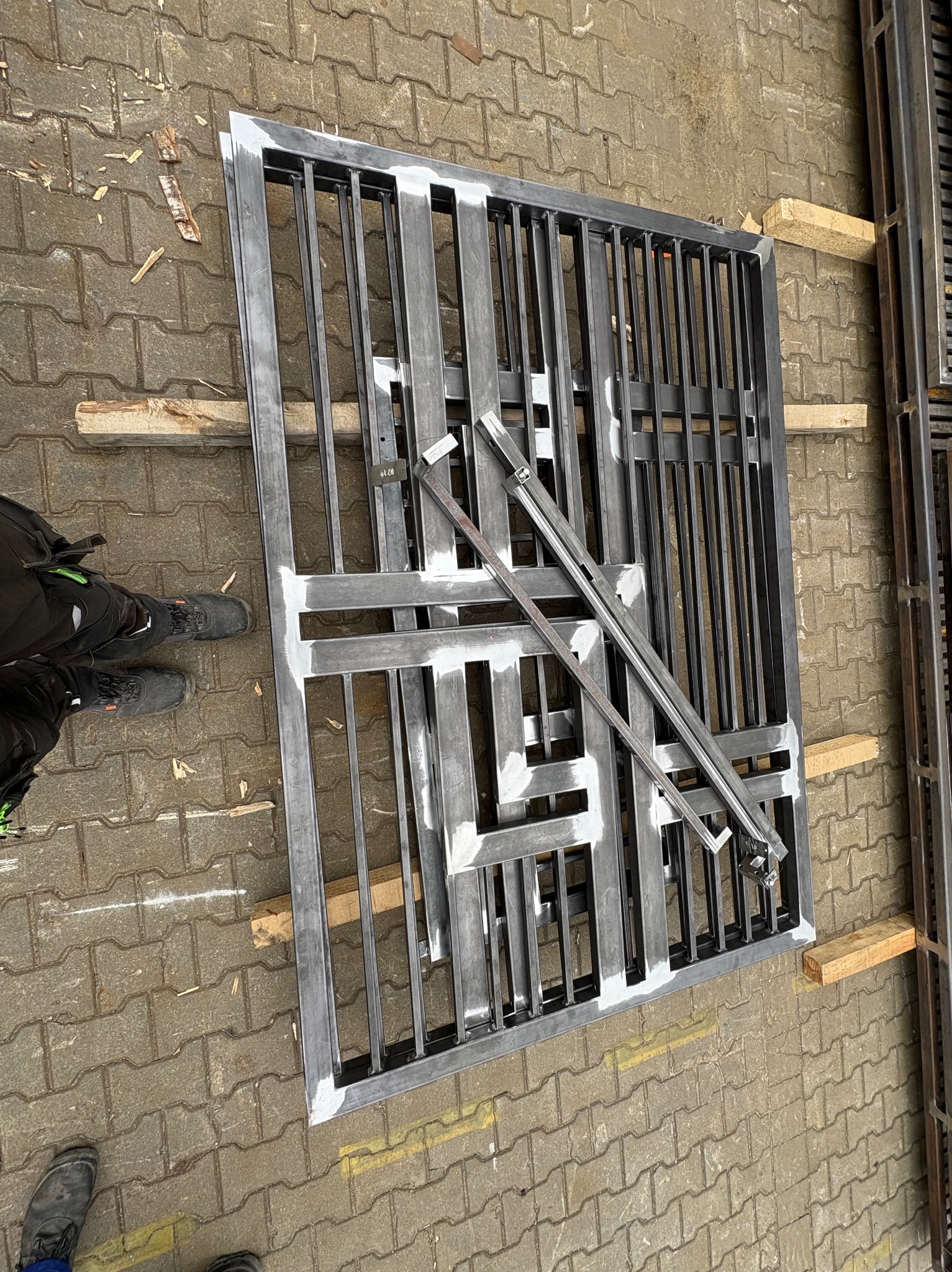 Metalowa konstrukcja kraty, prawdopodobnie fragment bramy lub ogrodzenia, leżąca na kostce brukowej. Widoczne ślady spawania i obróbki. Elementy wsparte na drewnianych klockach.