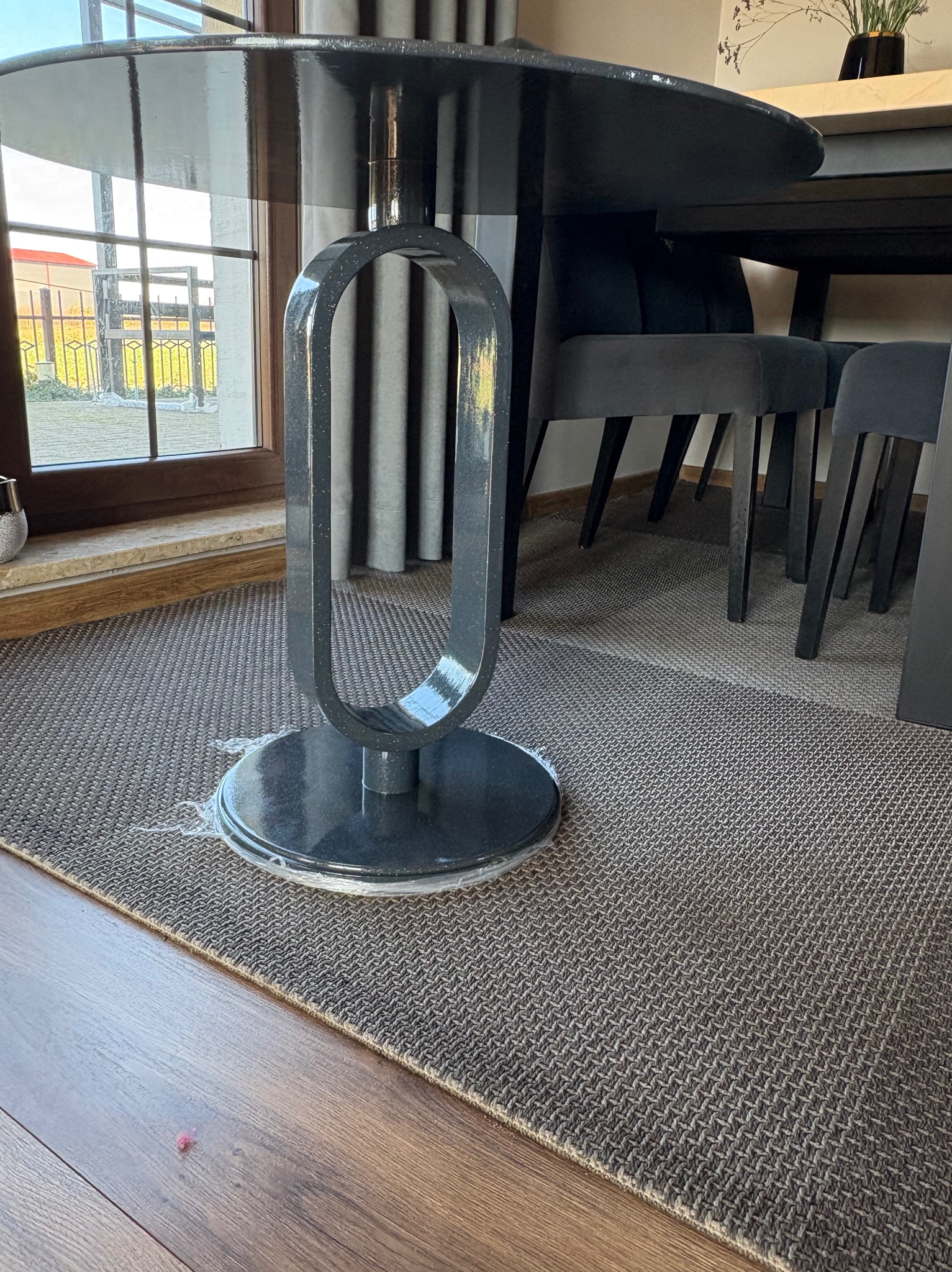 Stół z czarnym, błyszczącym blatem i oryginalną, metalową nogą w kształcie owalu. W tle krzesła i okno. Widoczny fragment dywanu i podłogi.