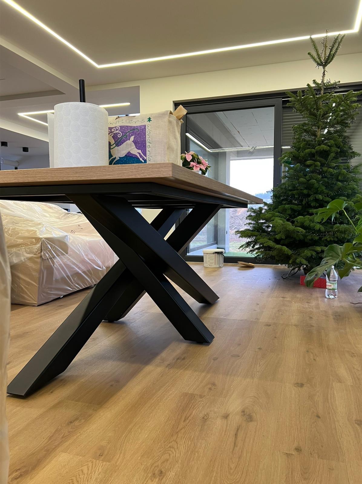 Stół z drewnianym blatem i czarnymi, metalowymi nogami w kształcie litery X. W tle okno i choinka. Widok z niskiej perspektywy, podkreślający konstrukcję stołu.