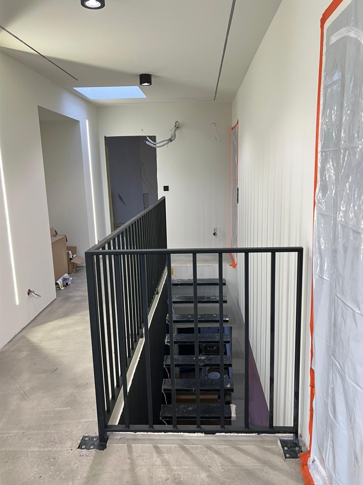 Nowoczesne schody metalowe z czarną balustradą w surowym wnętrzu. Widoczne przygotowania do dalszych prac wykończeniowych, zabezpieczone ściany folią malarską.