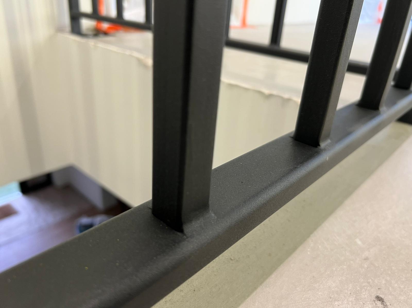 Czarne, metalowe balustrady schodowe z bliska, widoczne pionowe i poziome elementy konstrukcji. W tle jasne ściany i schody w dół. Minimalistyczny design.