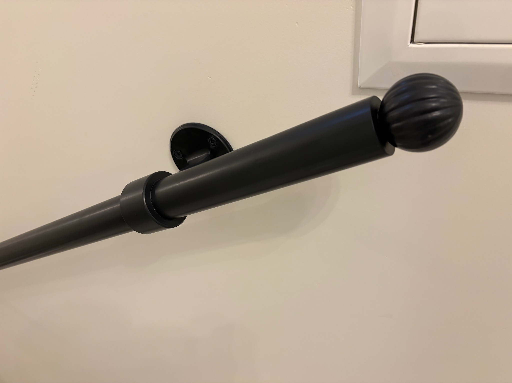 Czarna, metalowa poręcz z ozdobnym zakończeniem kulistym, zamocowana do jasnej ściany. Widoczne mocowanie do ściany i fragment rury. Minimalistyczny design.