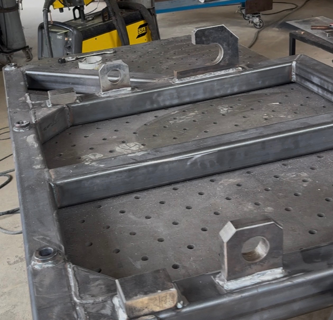 Metalowa konstrukcja spawana na perforowanym stole warsztatowym, widoczne spoiny i uchwyty montażowe. W tle spawarka ESAB.