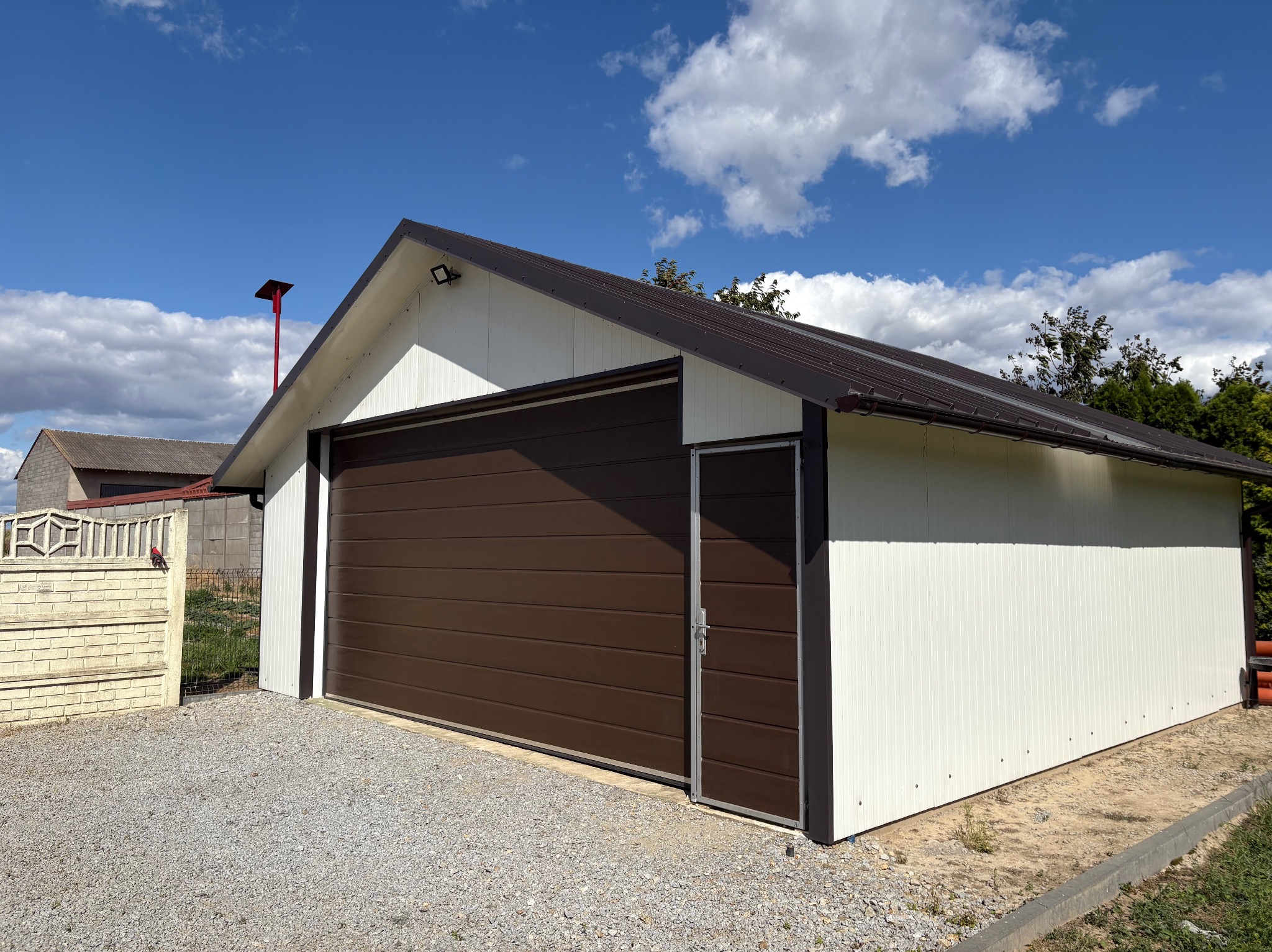 Garaż blaszany z brązową bramą segmentową i drzwiami wejściowymi, białe ściany z pionowym żłobkowaniem, na żwirowym podłożu, błękitne niebo z chmurami w tle.