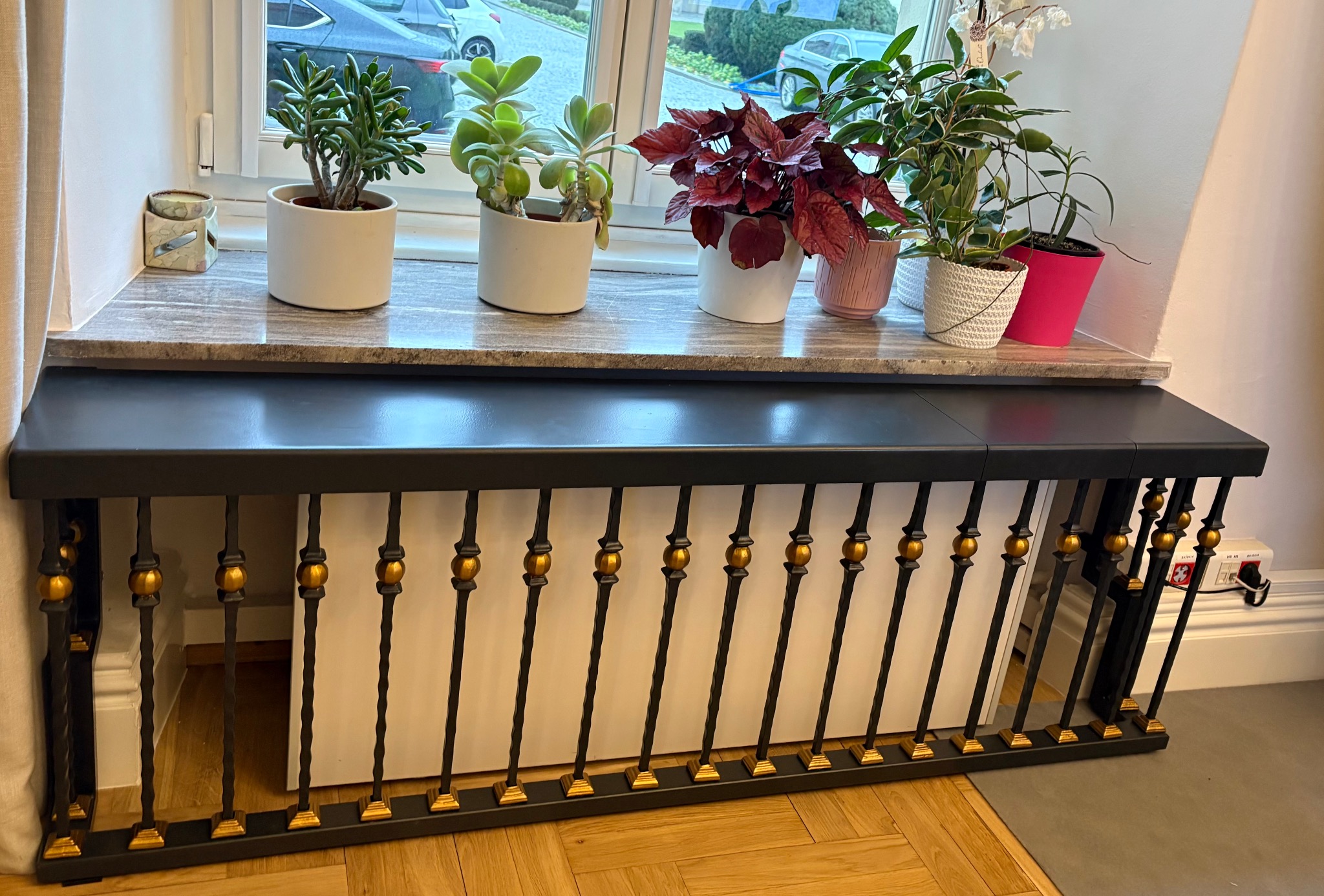 Dekoracyjny grzejnik z metalową balustradą w kolorze czarnym i złotym, na parapecie okna ustawione doniczki z roślinami. Nietypowe rozwiązanie aranżacyjne.