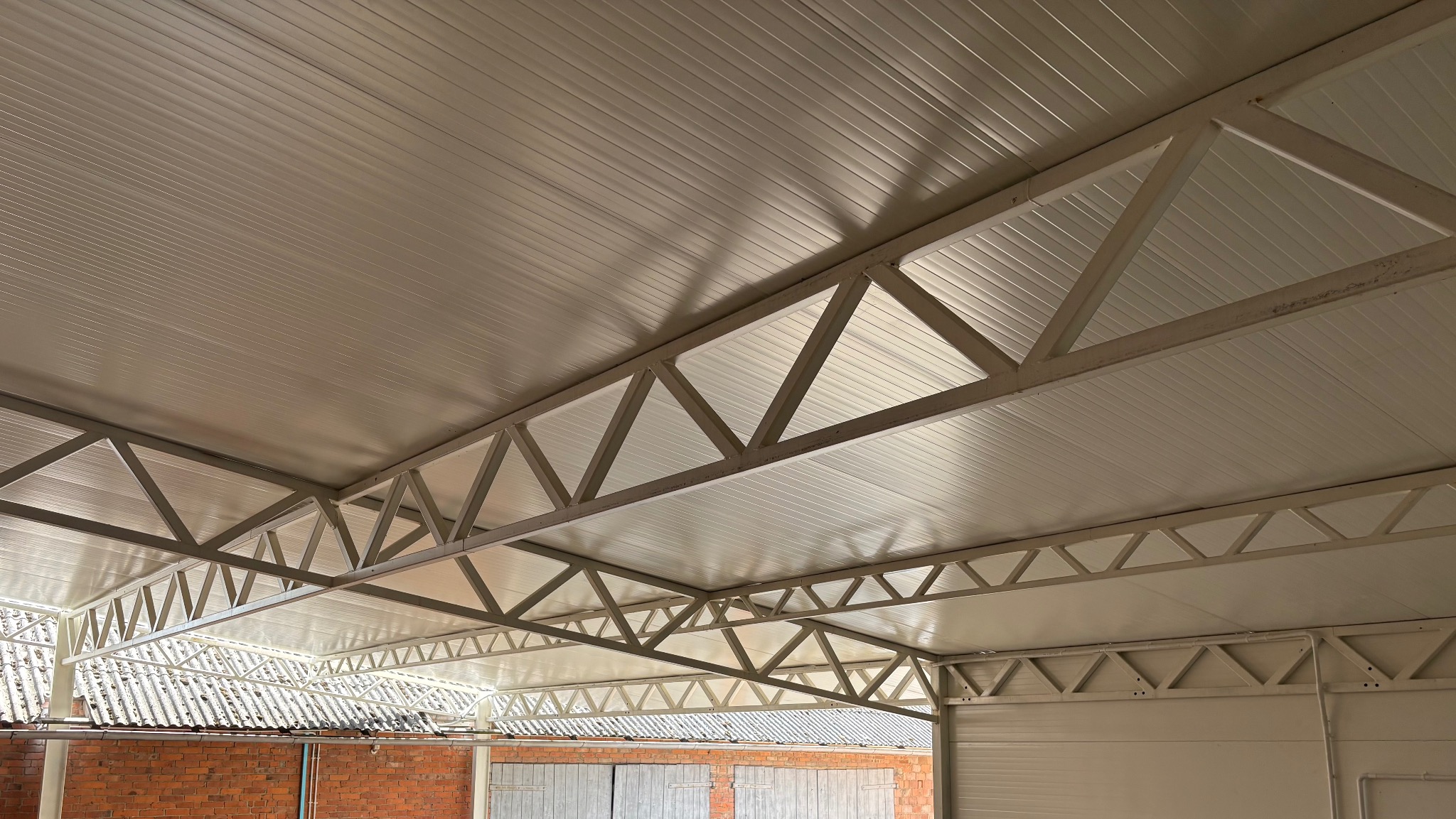 Metalowa konstrukcja zadaszenia garażu z blachy trapezowej, widok od wewnątrz, z fragmentem ceglanego budynku w tle. Jasne oświetlenie.