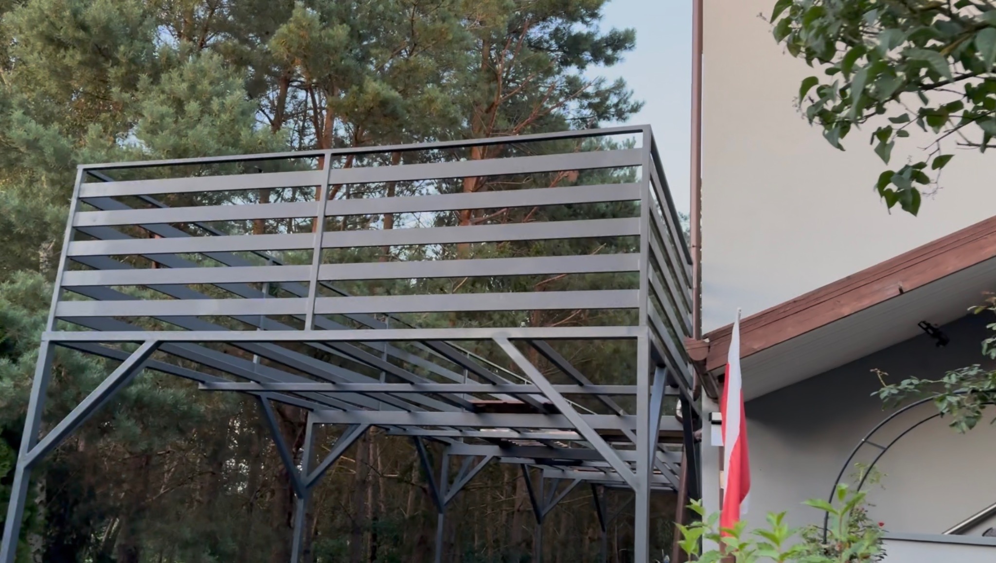 Metalowa konstrukcja z poziomymi belkami, prawdopodobnie zadaszenie tarasu, na tle drzew i fragmentu budynku z polską flagą. Szara, minimalistyczna forma.
