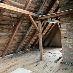 Stare poddasze z drewnianą konstrukcją dachu, ceglanym kominem i widocznymi śladami zniszczeń, przygotowane do opróżnienia lub remontu.