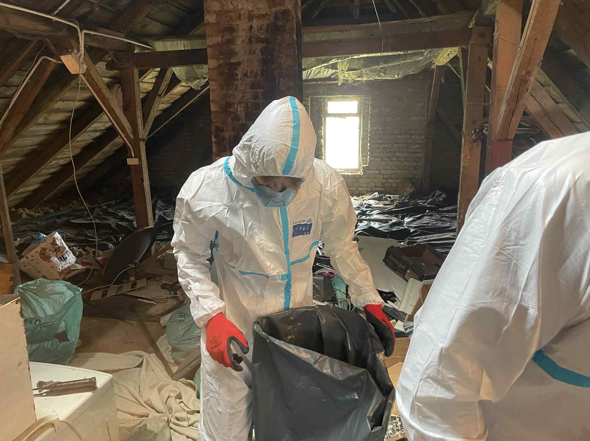 Pracownik w kombinezonie ochronnym i czerwonych rękawicach zbiera śmieci do worka na strychu z drewnianą konstrukcją i oknem w tle. Widoczne porozrzucane przedmioty.