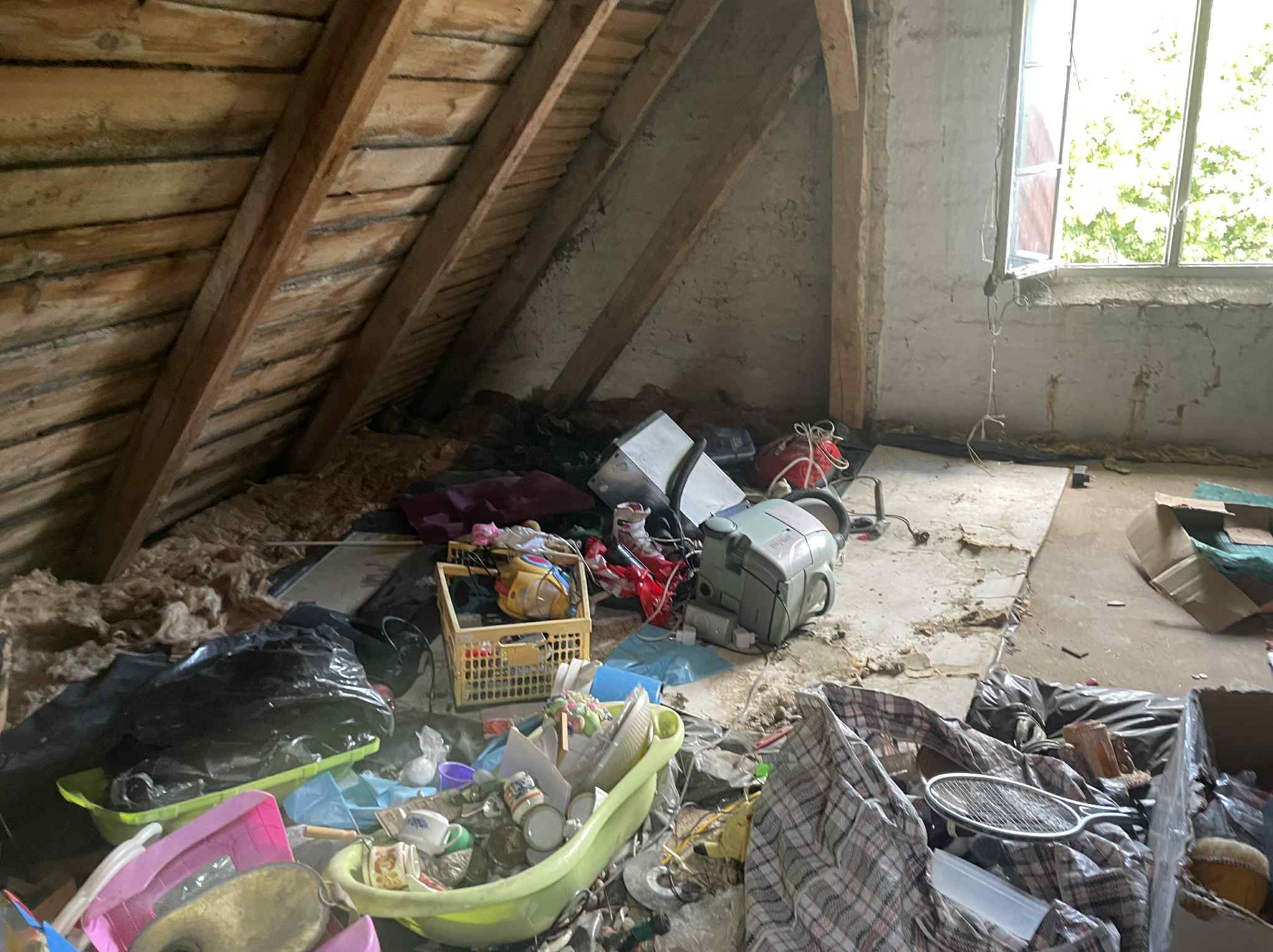 Zagracone poddasze ze skosami i oknem. Różne przedmioty w pojemnikach i workach, w tym zabawki, naczynia i sprzęt AGD. Widoczne drewniane elementy konstrukcyjne dachu.