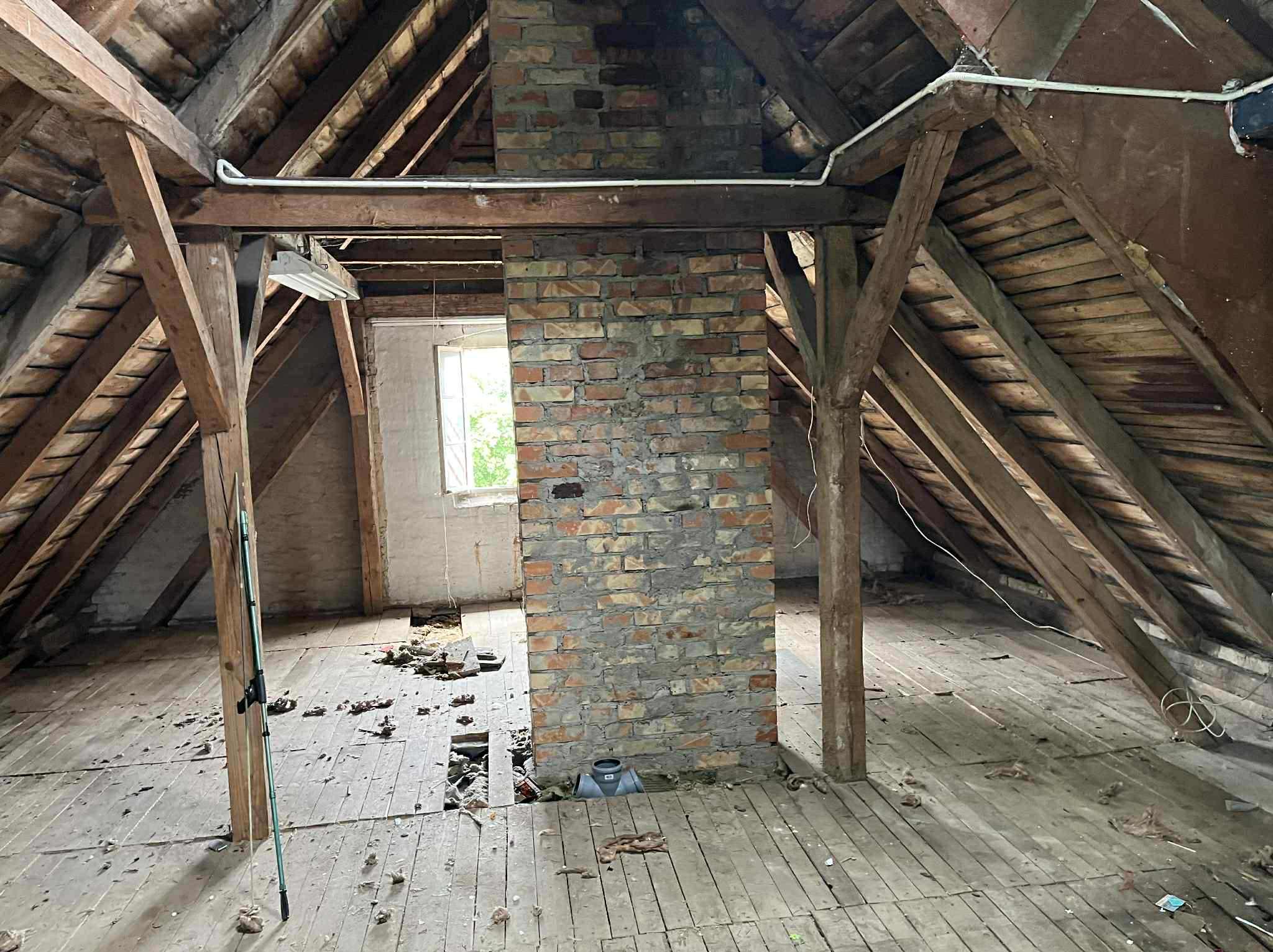 Strych z odsłoniętą konstrukcją dachu i kominem z cegły, widoczne ślady po demontażu, resztki materiałów na podłodze, okno w tle. Widok na poddasze.