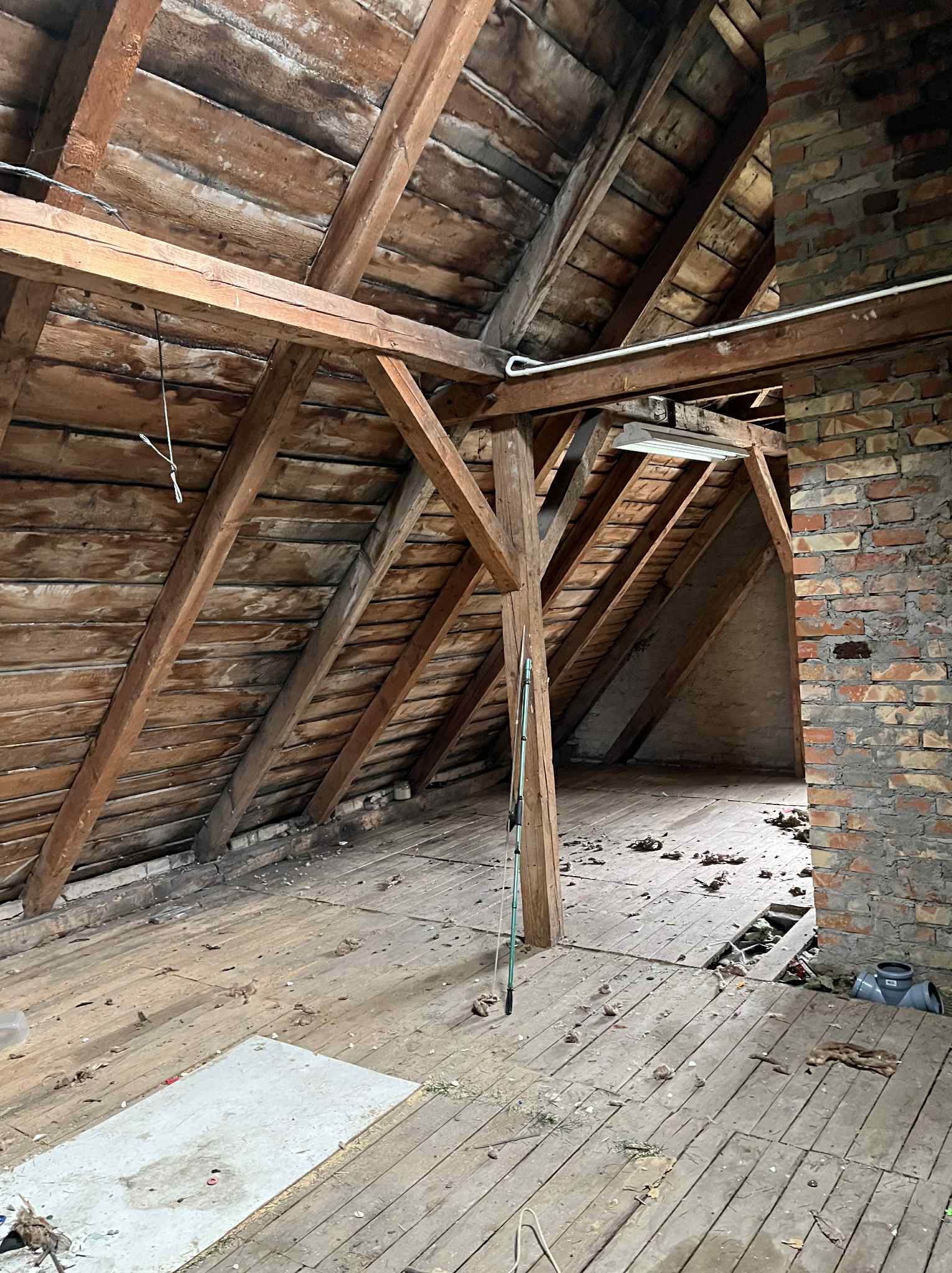 Stare poddasze z drewnianą konstrukcją dachu, ceglanym kominem i widocznymi śladami zniszczeń, przygotowane do opróżnienia lub remontu.