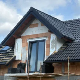 Częściowy widok domu w trakcie krycia dachu czarną dachówką. Widoczne rusztowania, okno dachowe i nieotynkowane ściany z pustaków.