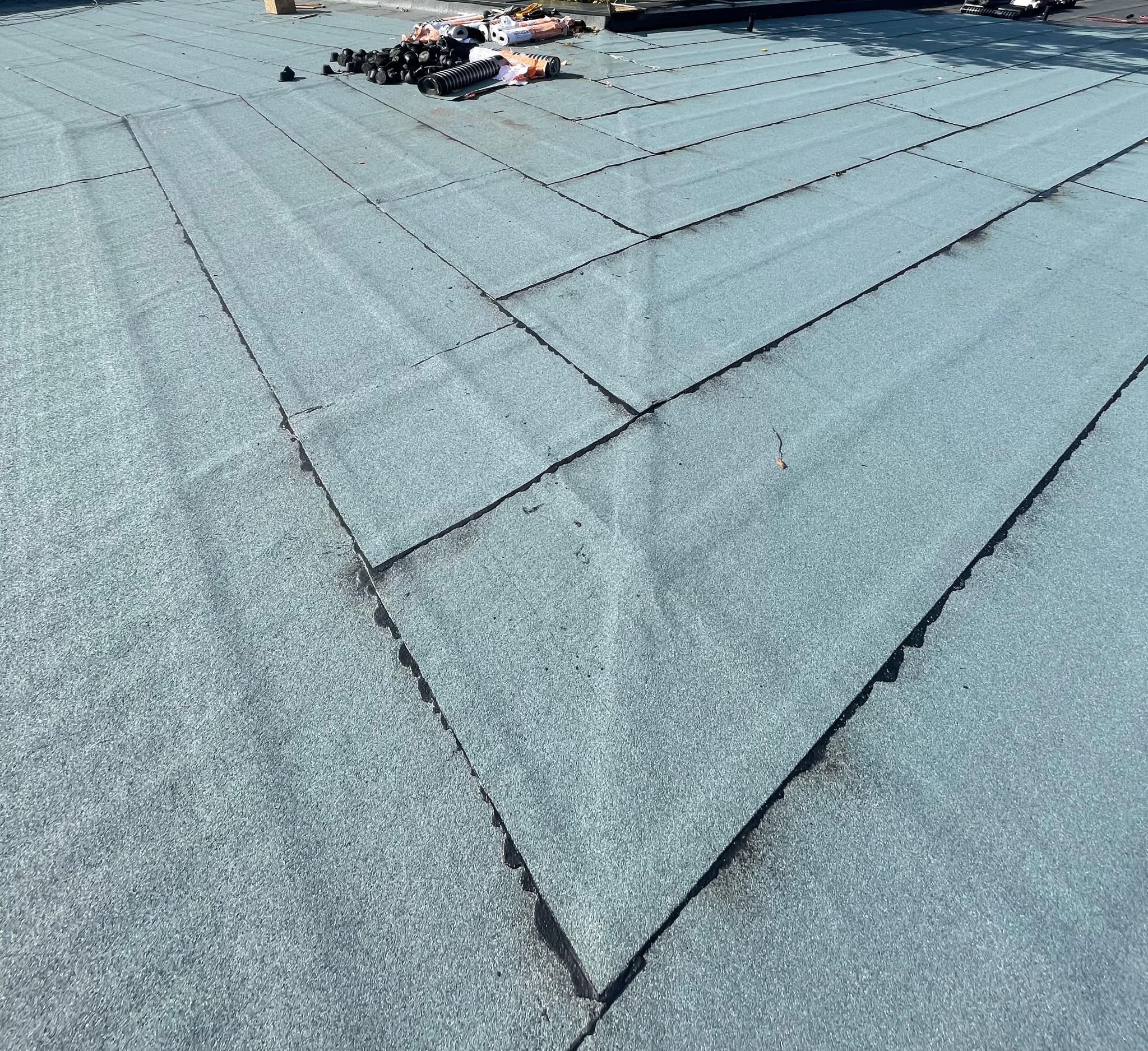 Widok z góry na płaski dach pokryty papą termozgrzewalną, z widocznymi łączeniami i narzędziami dekarskimi w tle. Dach w odcieniach szarości i błękitu.