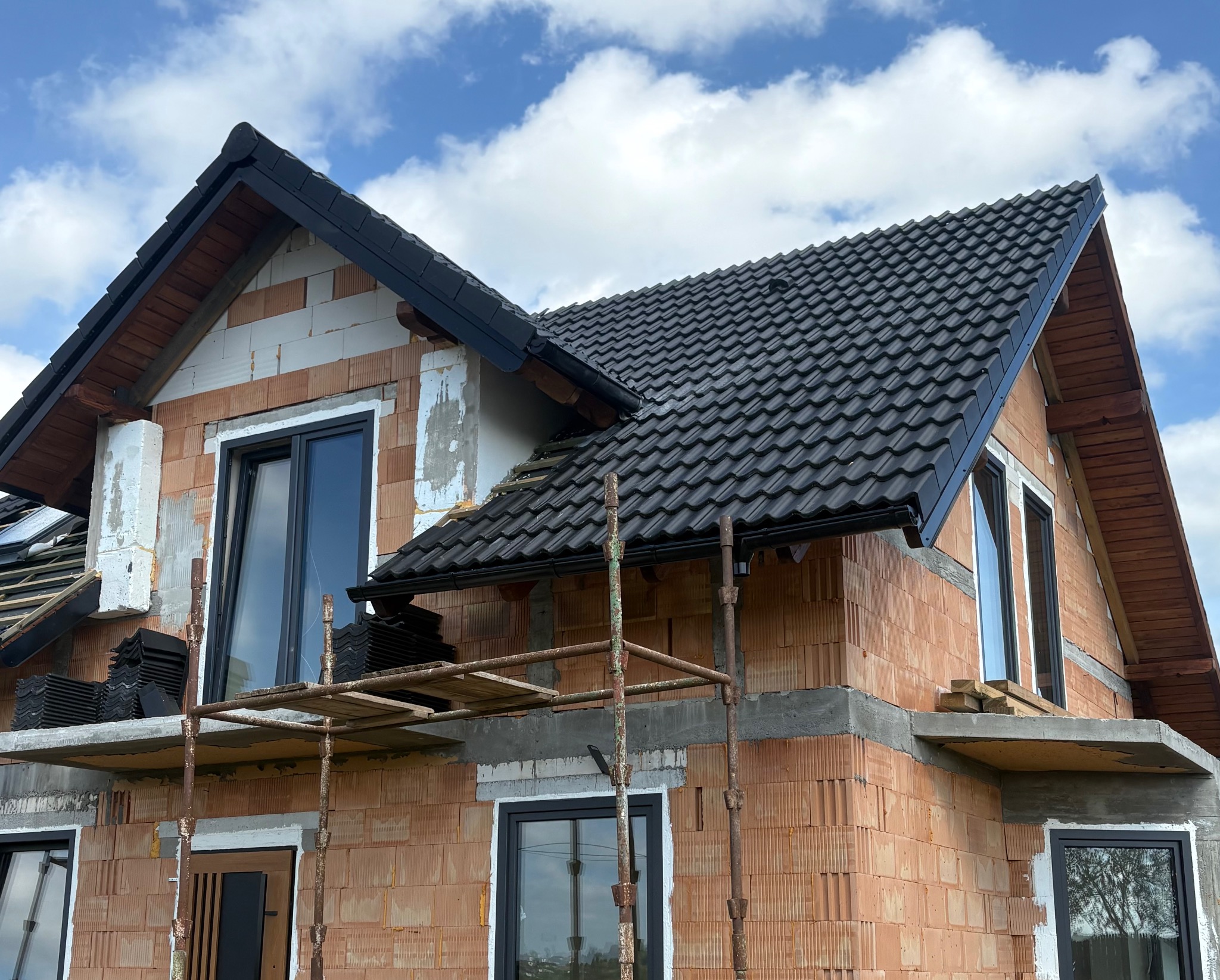 Dom w trakcie budowy z czarną dachówką i rynnami. Widoczne rusztowania i niezakończone obróbki. Częściowo pokryty dach na tle błękitnego nieba z chmurami.