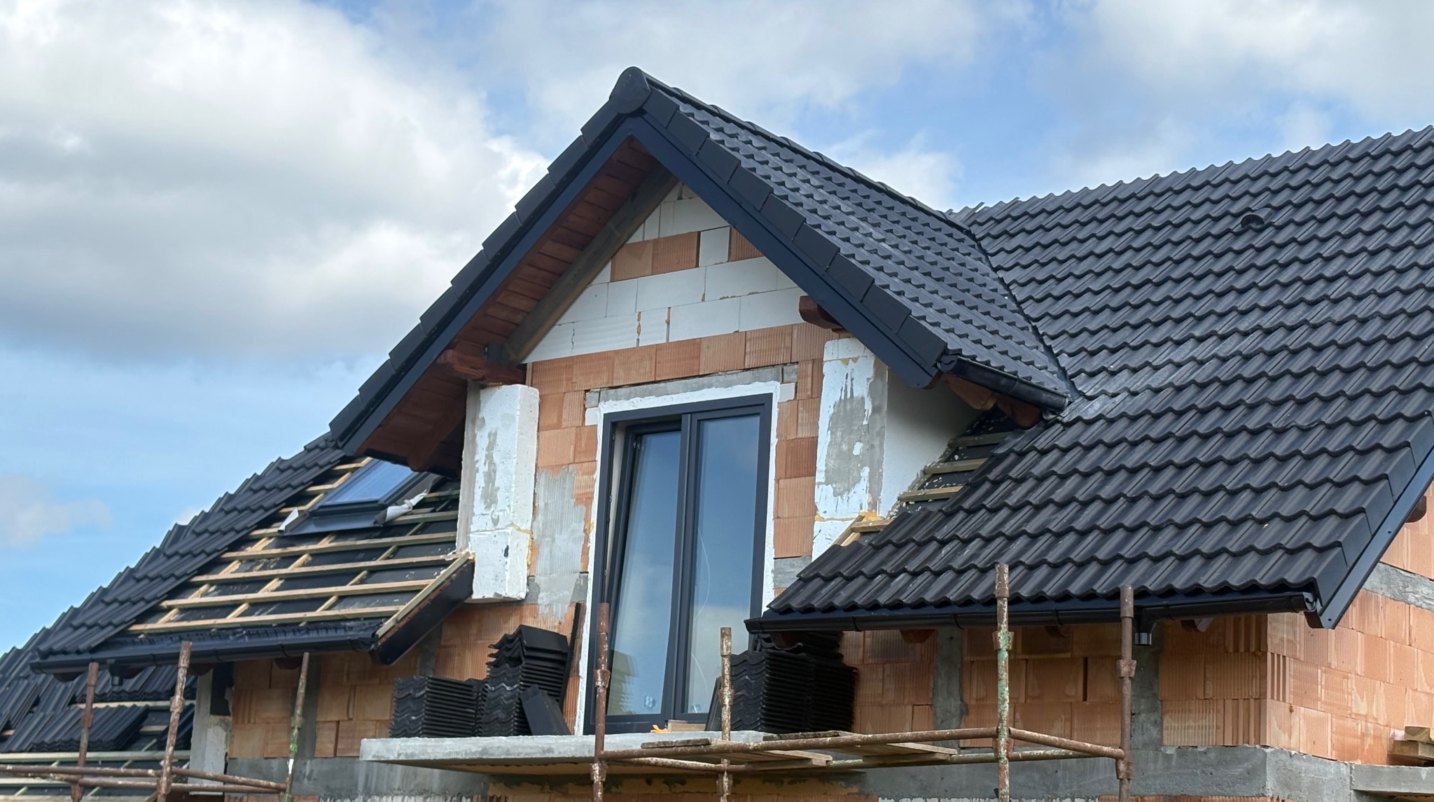 Częściowy widok domu w trakcie krycia dachu czarną dachówką. Widoczne rusztowania, okno dachowe i nieotynkowane ściany z pustaków.