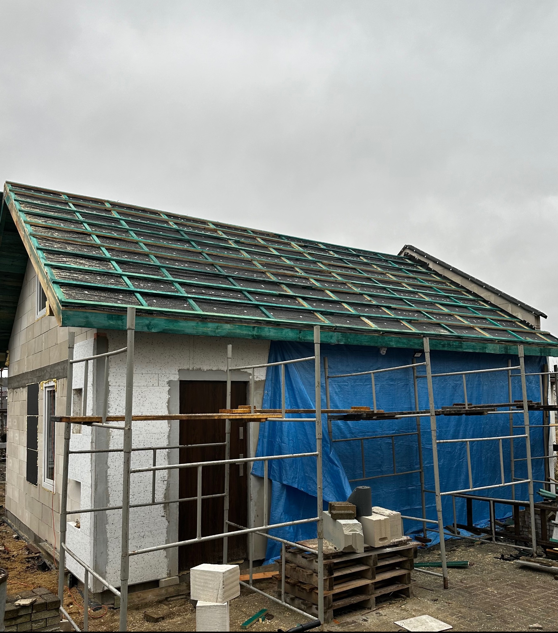 Dom w trakcie budowy, widoczne rusztowanie i dach z łatami pod pokrycie. Na dachu papa. Ściany z białego bloczka. Całość przykryta niebieską plandeką.