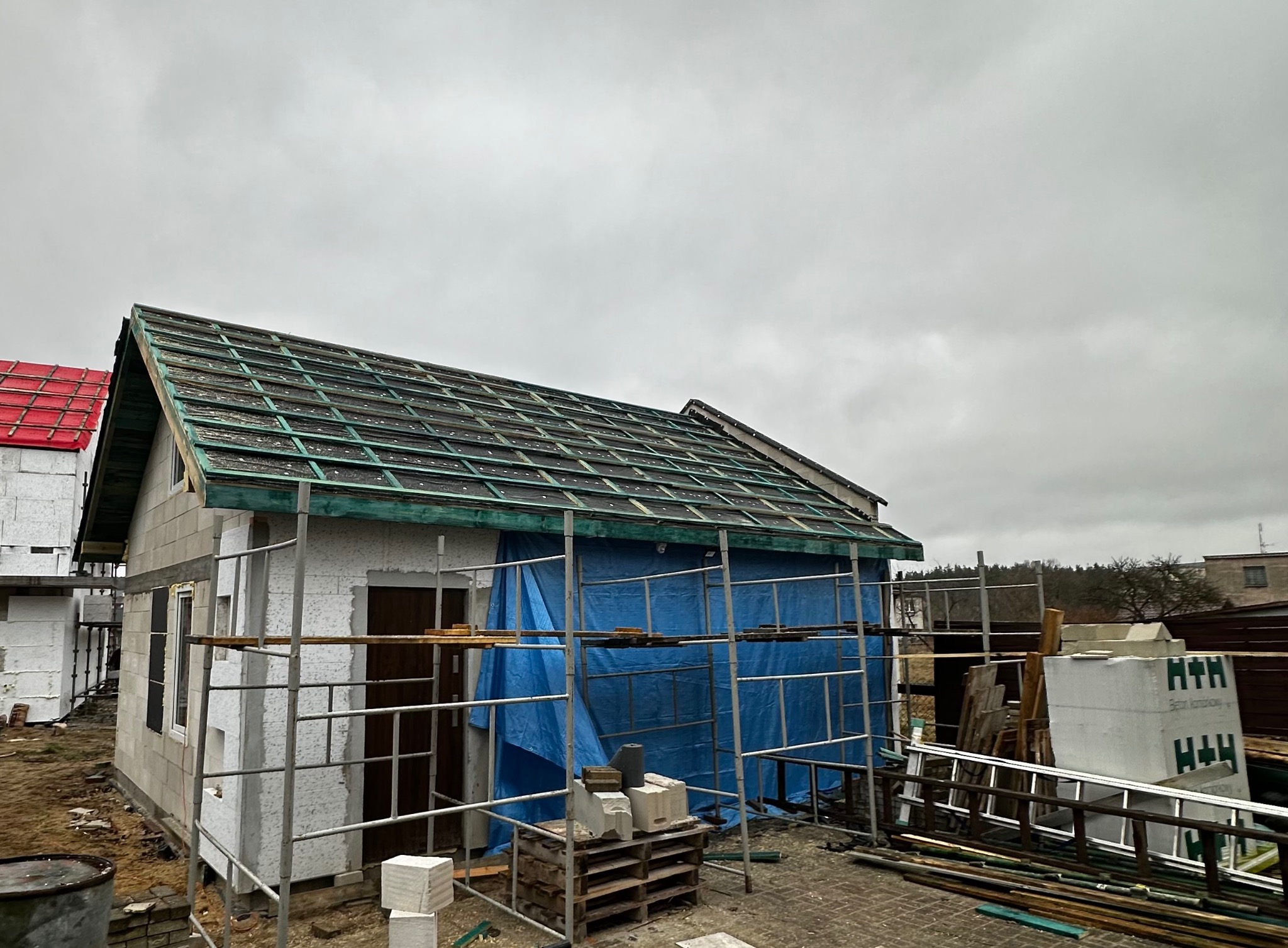 Dom w trakcie budowy z częściowo pokrytym dachem, zabezpieczony folią i otoczony rusztowaniami. Widoczne materiały budowlane i elementy konstrukcyjne.