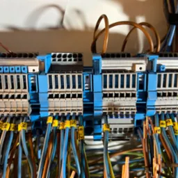 Szczegółowe ujęcie rozdzielnicy elektrycznej z oznaczonymi przewodami, widoczne niebieskie i szare elementy złączek. Przewody w różnych kolorach z żółtymi oznaczeniami.