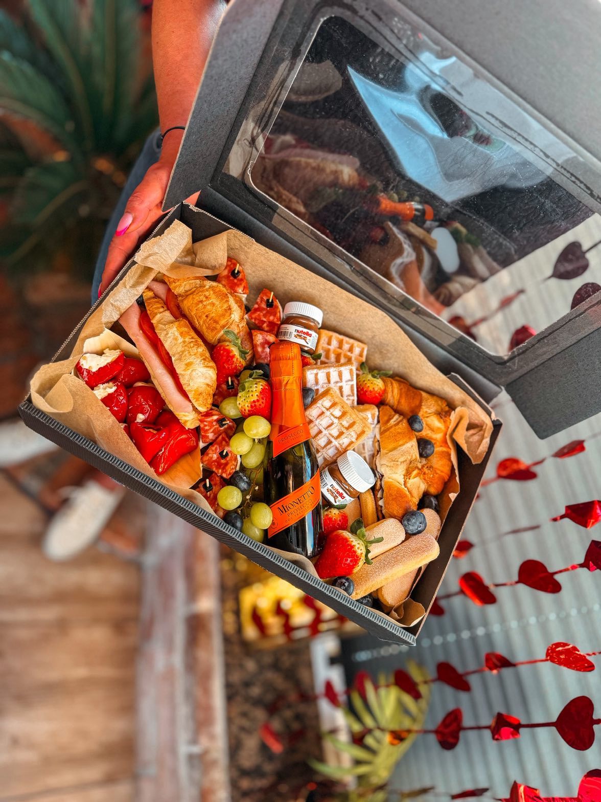 Otwarta, czarna skrzynka z przezroczystym wieczkiem wypełniona przekąskami: croissantami, goframi, owocami, serami i butelką Prosecco. Romantyczna dekoracja w tle.