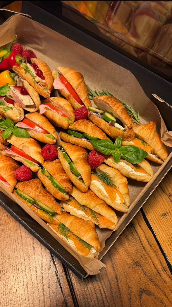 Elegancka taca z mini croissantami z różnymi nadzieniami, udekorowana malinami i ziołami, prezentowana na drewnianym stole. Idealne na catering.