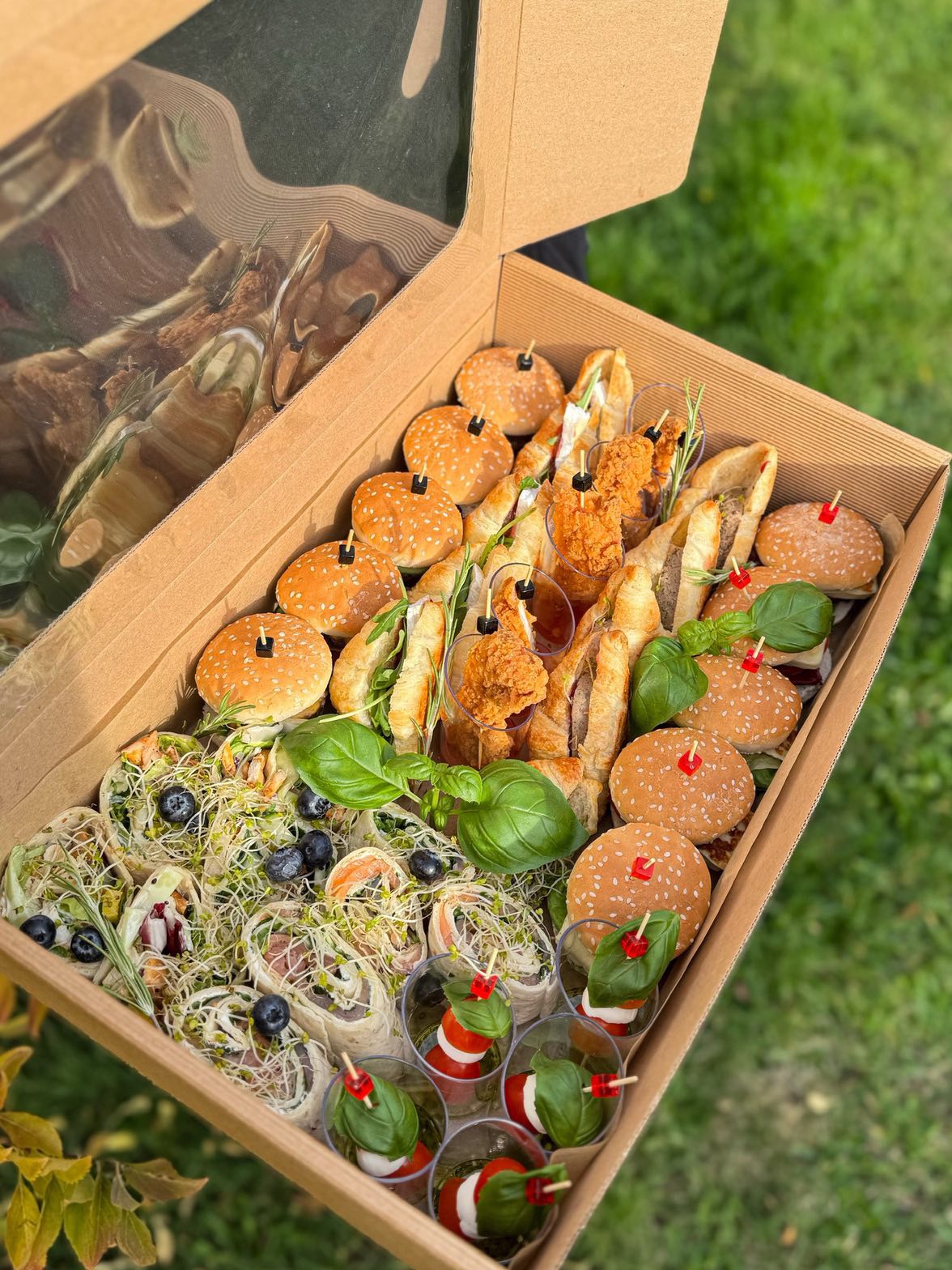 Różnorodna przekąska w kartonowym pudełku z przezroczystym wieczkiem: mini burgery, tortille, roladki, mini sałatki caprese, smażony kurczak, dekoracje z bazylii i rozmarynu.