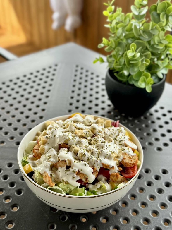Apetyczna sałatka z kurczakiem, grzankami i sosem, posypana ziołami, w białej misce na tle szarego stołu i zielonej roślinki.