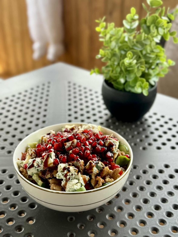 Apetyczna sałatka z serem pleśniowym, orzechami i granatem w białej misce na tle doniczki z zieloną rośliną, ustawiona na metalowym stole.