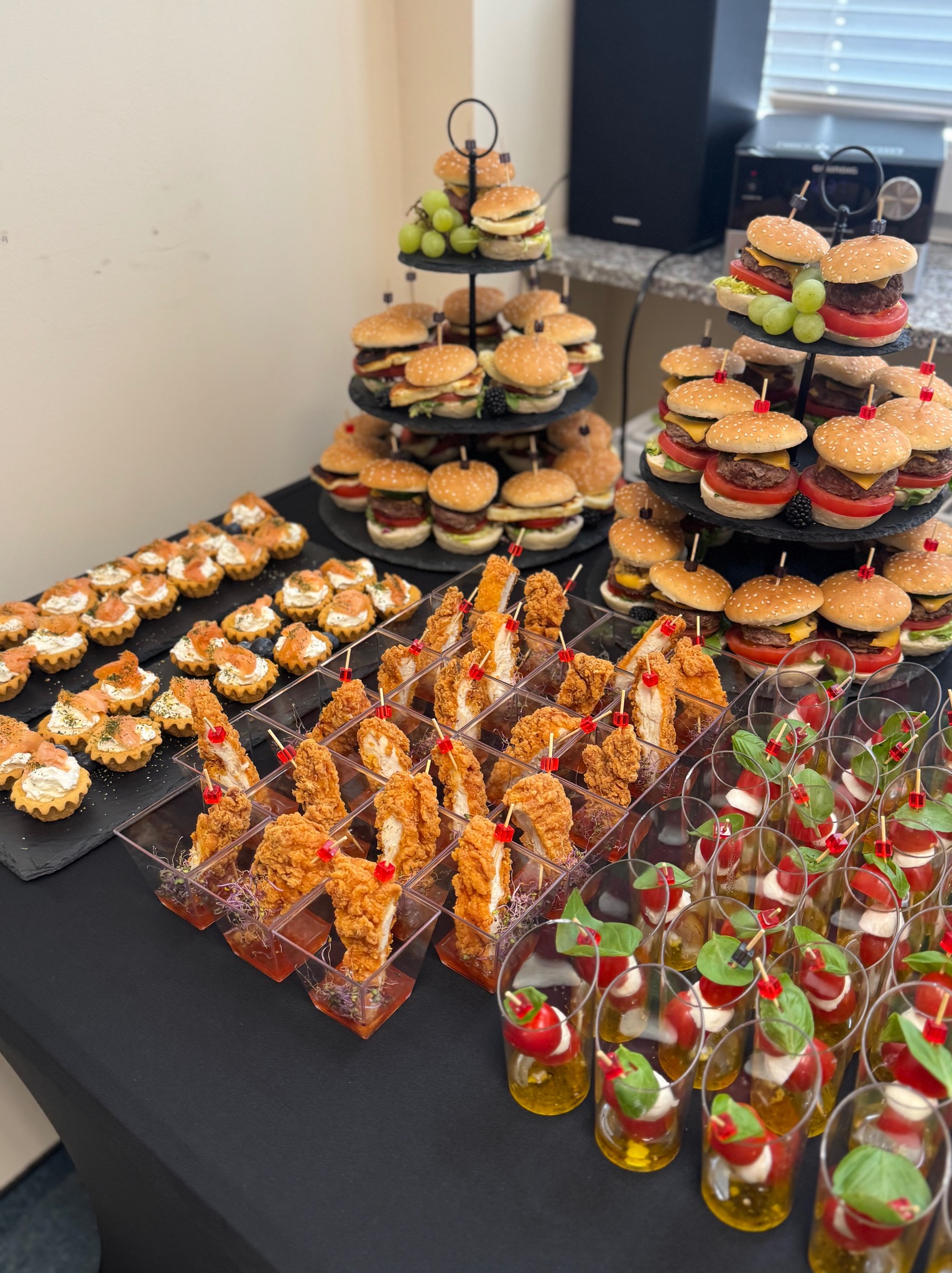 Stół z różnorodnymi przekąskami: mini burgery na paterach, tartaletki z łososiem, kurczak w panierce w kieliszkach i sałatka caprese w małych szklankach.