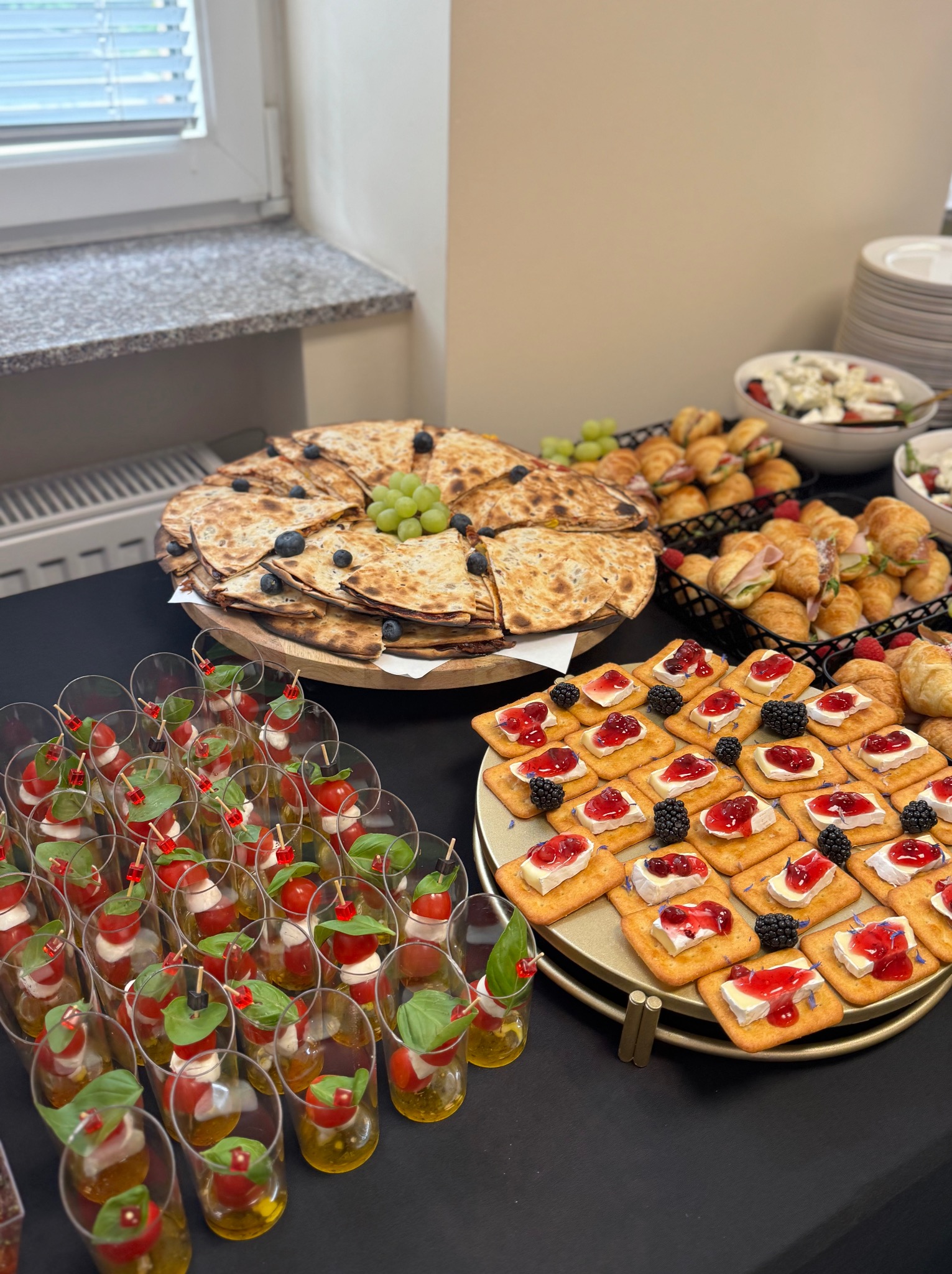 Elegancki stół cateringowy z przekąskami: mini-sałatki w szklankach, pieczywo z owocami, kanapki z serem i dżemem, croissanty. Aranżacja pod światłem okna.