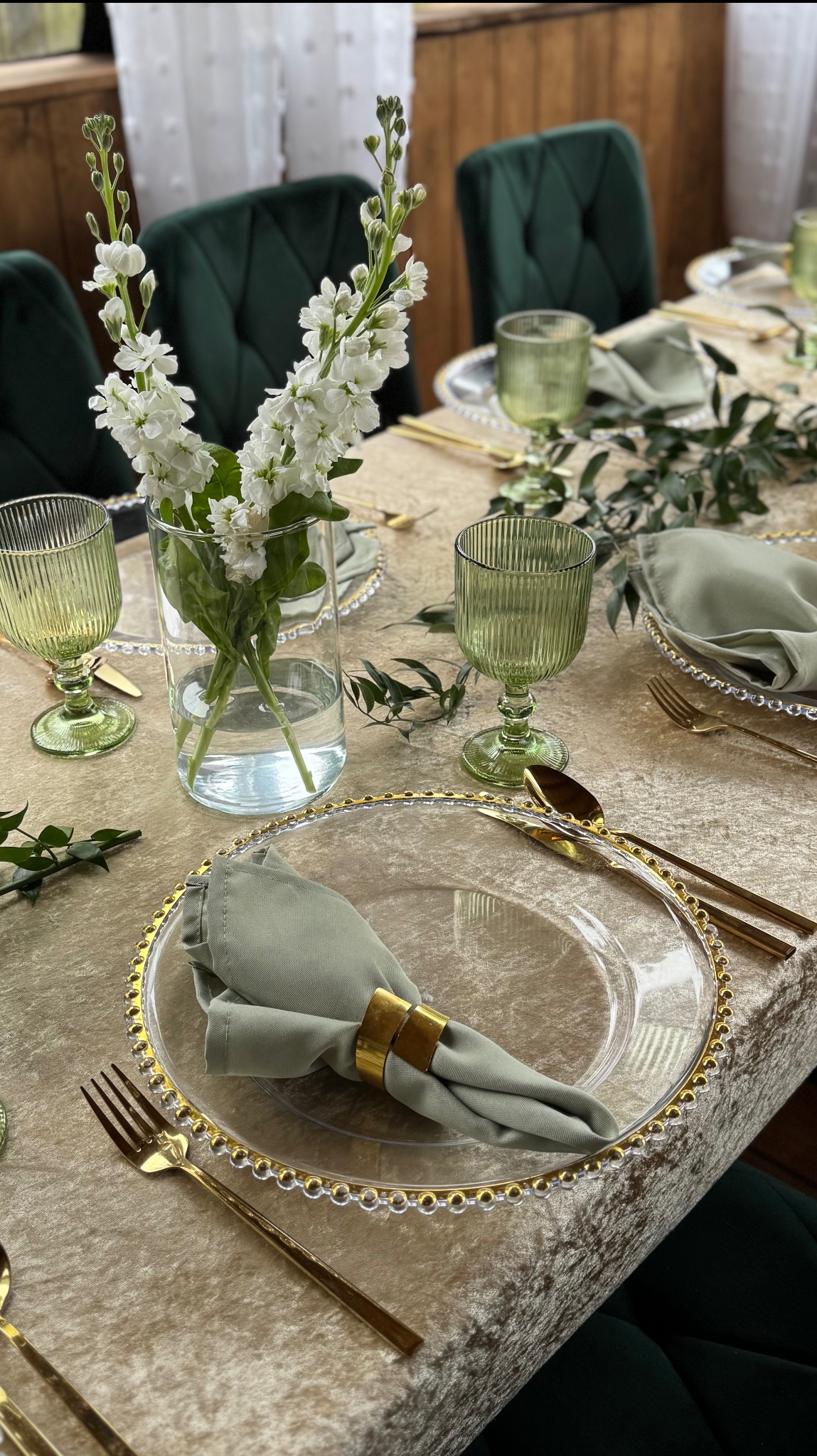 Elegancka aranżacja stołu: złote sztućce, szklane talerze z obwódką, zielone serwetki i kieliszki, kwiaty w wazonie. Idealne na uroczystości.