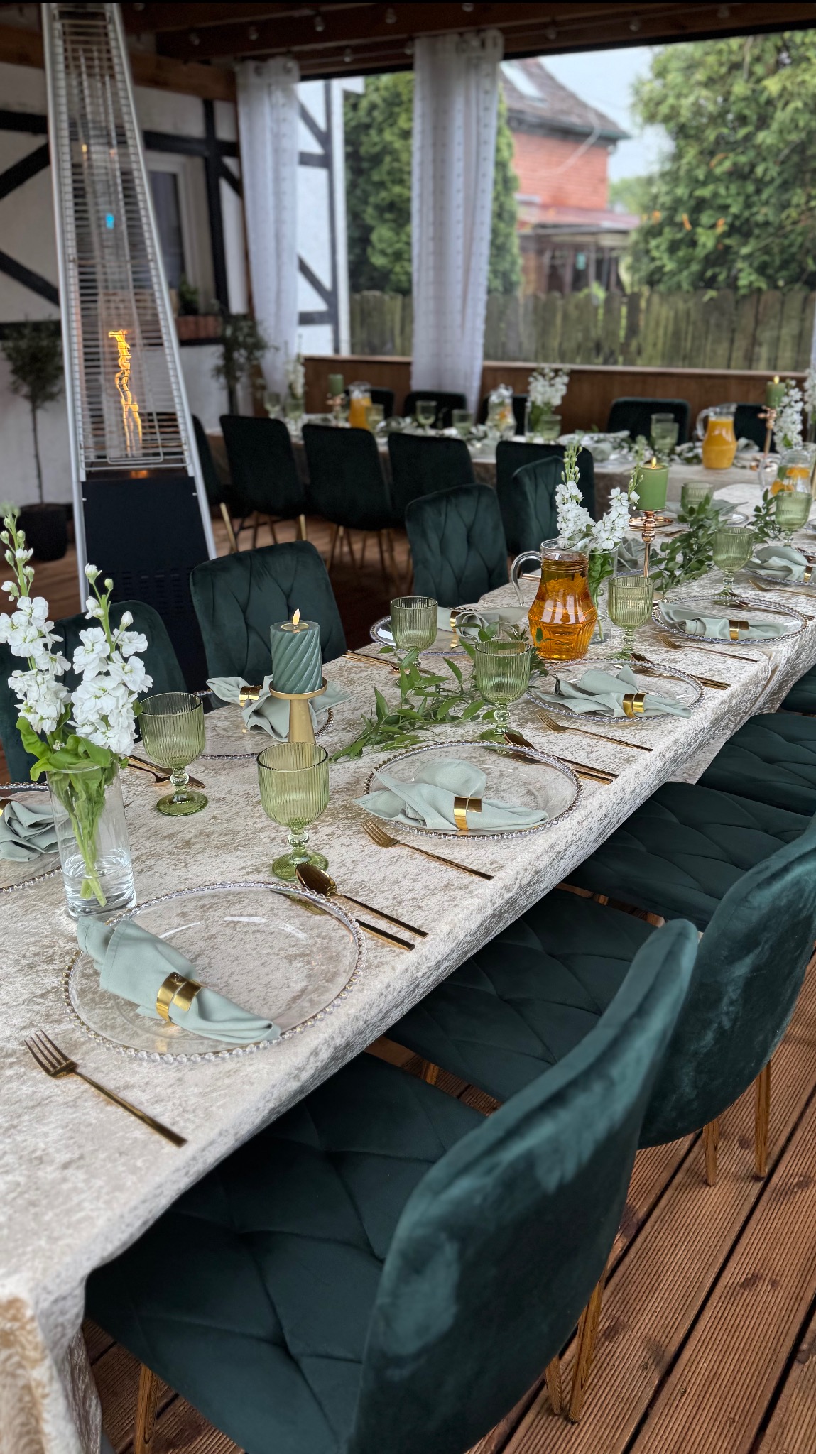 Długi stół z elegancką zastawą w odcieniach zieleni i złota, udekorowany kwiatami i świecami, ustawiony na zewnątrz, prawdopodobnie na przyjęcie.