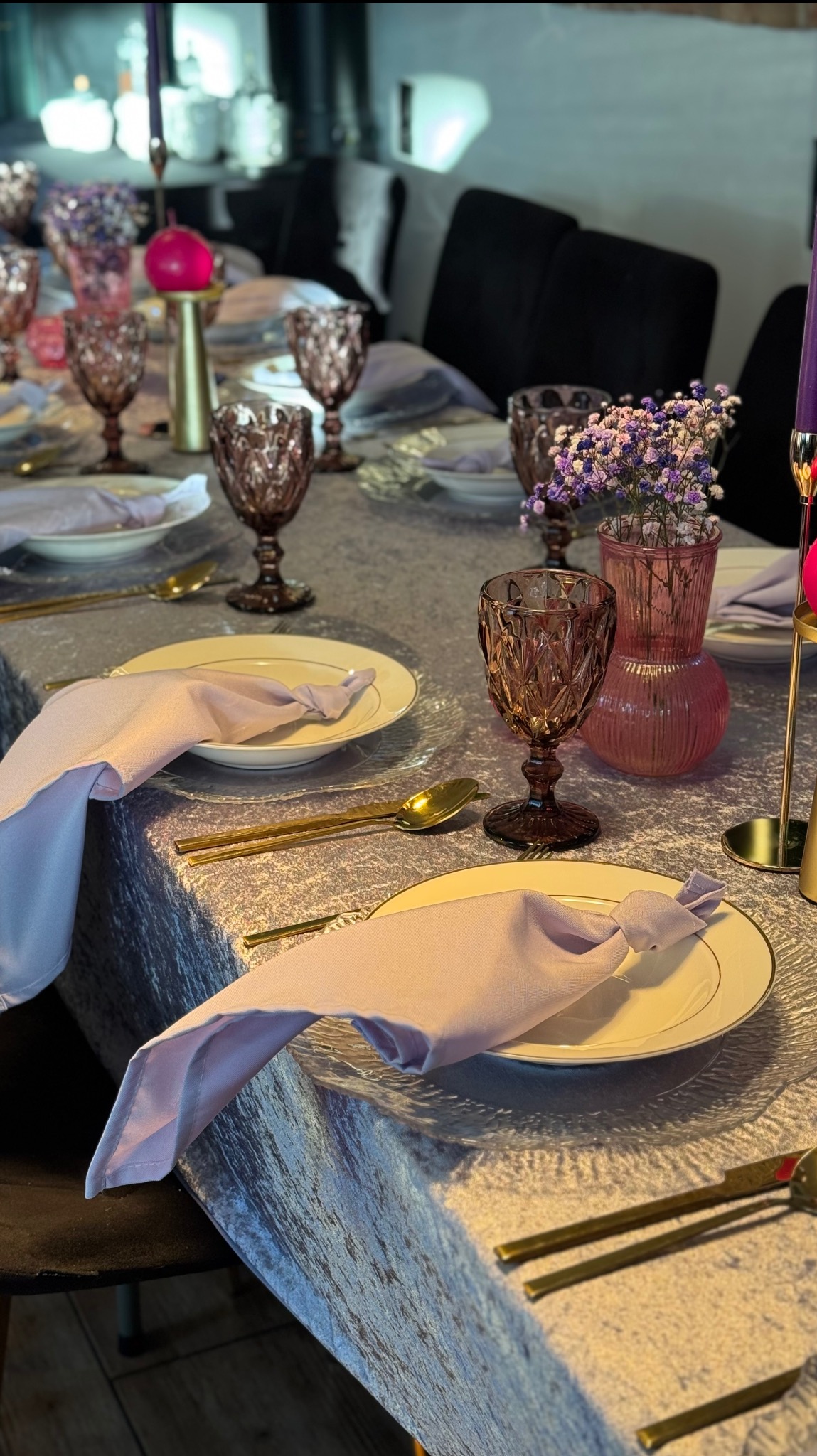 Elegancko nakryty stół z fioletowymi akcentami: kieliszki, serwetki, kwiaty w wazonach. Złote sztućce i świeczniki dodają blasku aranżacji.