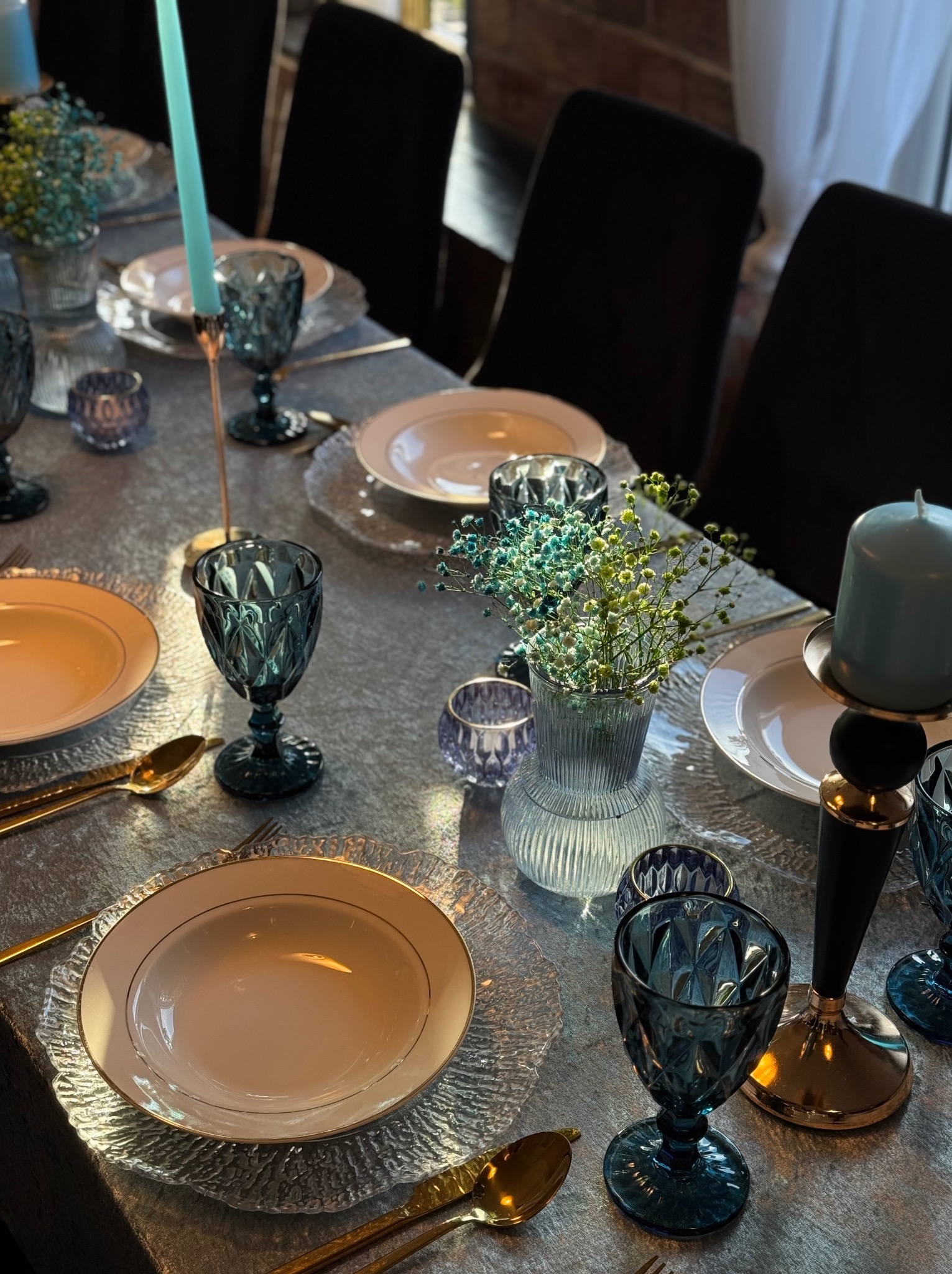 Elegancko nakryty stół z błękitnymi akcentami: kieliszki, świece i kwiaty. Dekoracja stołu z dbałością o detale, złote sztućce, teksturowane podkładki pod talerze.